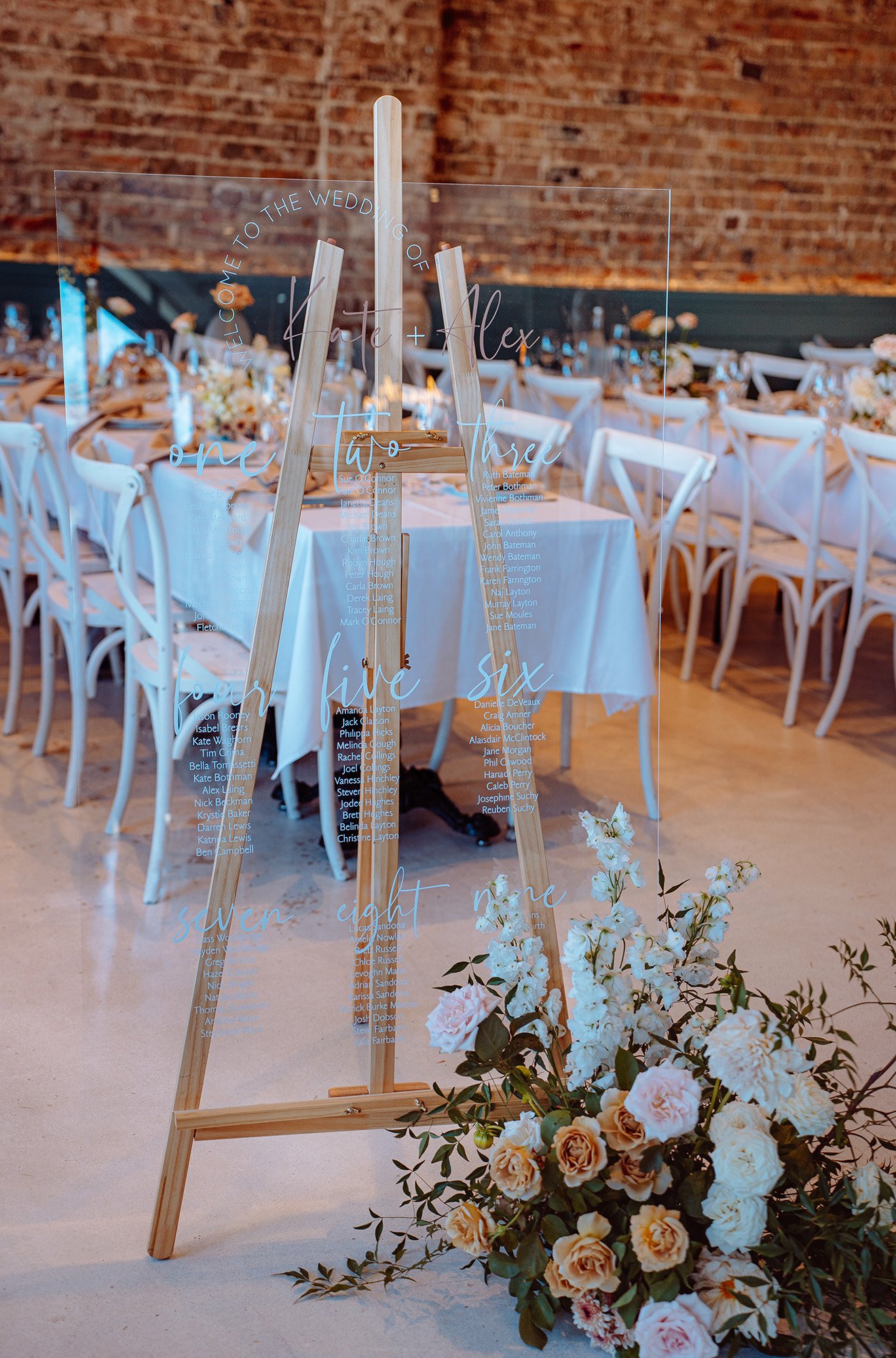 A wedding seating chart on a glass panel with a wooden stand, surrounded by white and blush flowers, set against a brick wall and dining tables with white chairs in a rustic venue.
