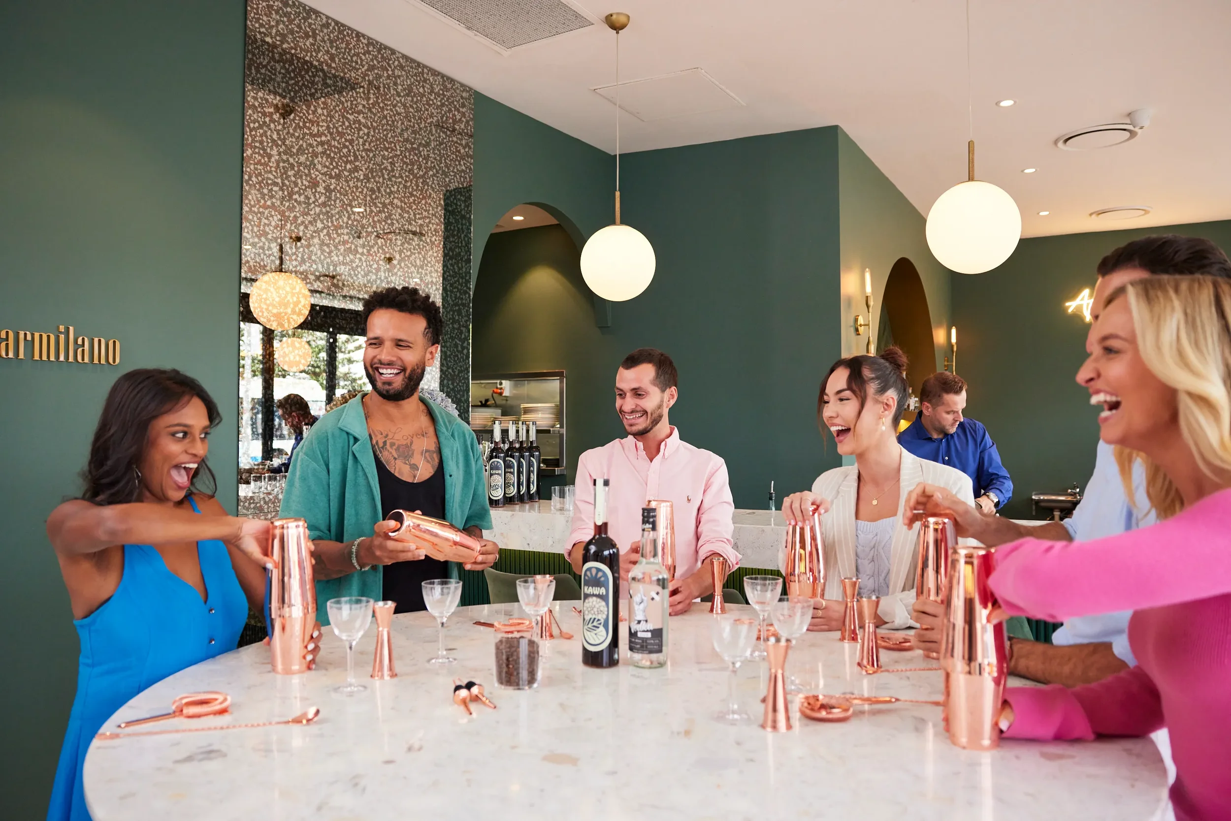A group of six diverse people smiling and enjoying cocktails at a bar or restaurant. The bar has a white marble counter and is decorated with bottles, copper cups, and glasses. The background shows green walls, modern lighting, and decorative mirrors