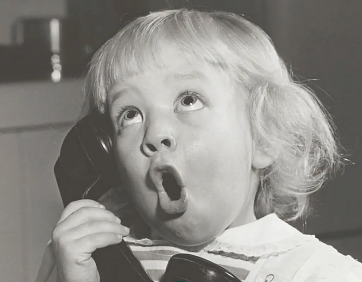 Black and white photo of a young girl with curly, light-colored hair talking on a corded phone, with her mouth open and eyes wide.
