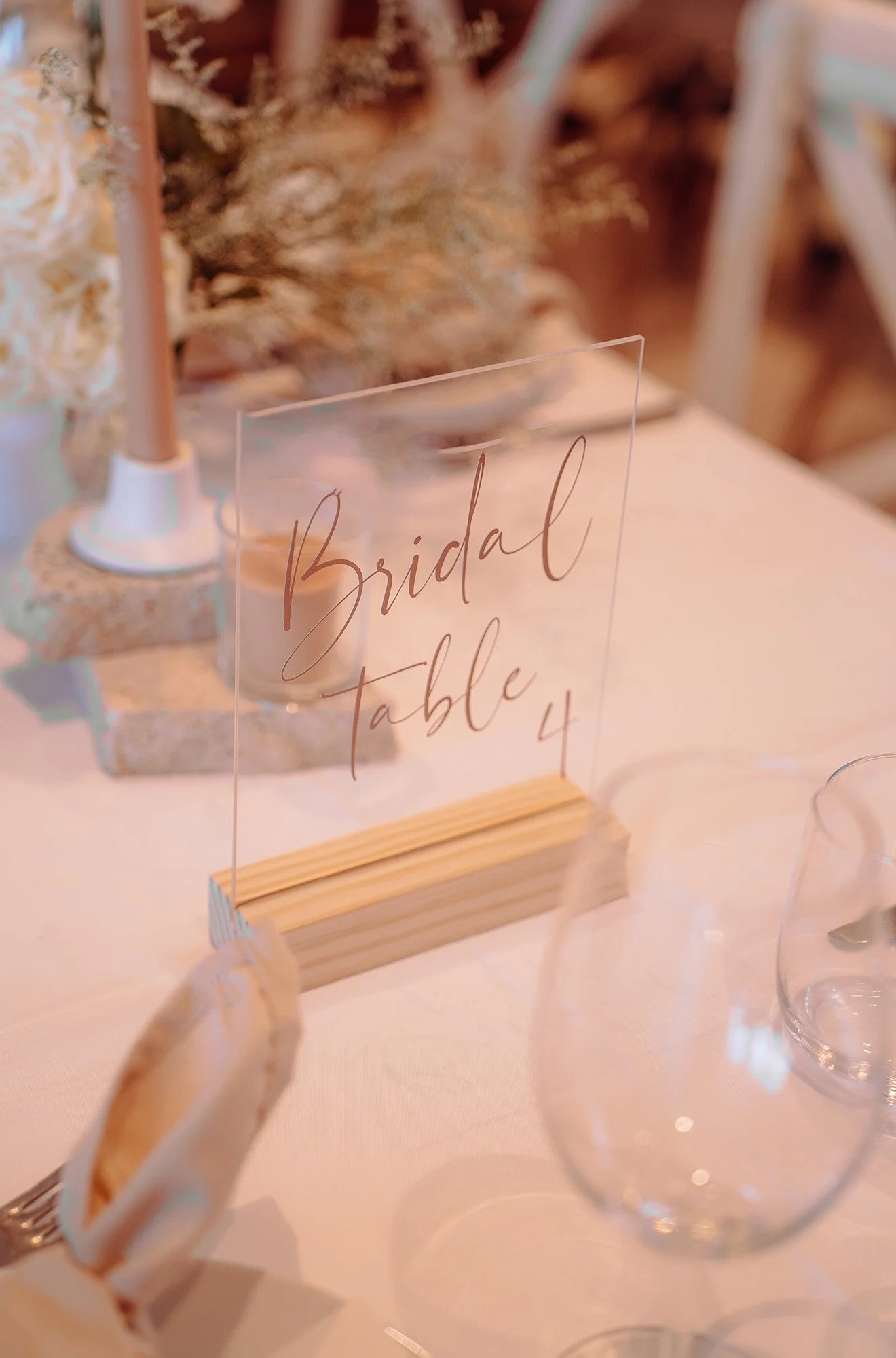 A clear acrylic sign with elegant handwritten text that reads 'Bridal table 4' is placed on a light-colored table, surrounded by a floral arrangement and glassware, indicating a wedding reception setting.
