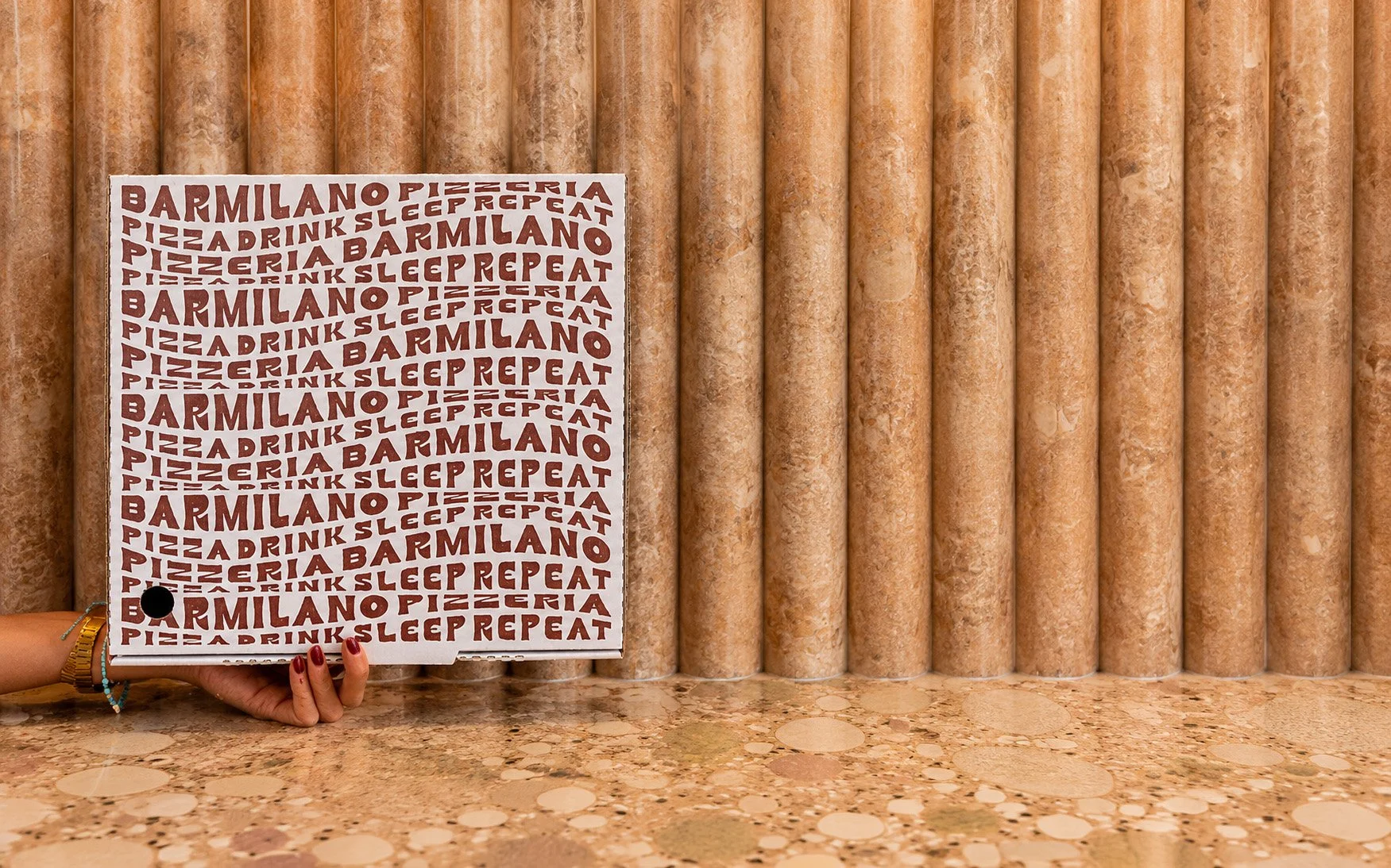 Person holding a black pen and wearing a gold bracelet with colorful beads, standing in front of a wall of large beige marble columns, holding a white sign with red text that reads 'BARMILANO PIZZERIA PIZZA DRINK SLEEP REPEAT'.