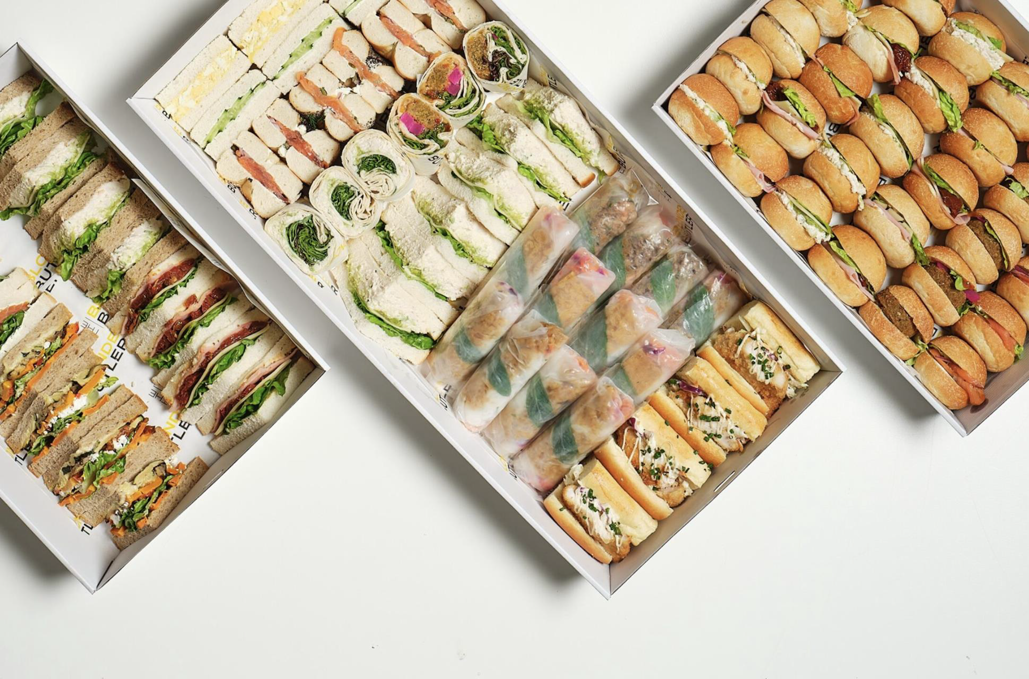Three open white boxes filled with assorted mini sandwiches and wraps, including varieties with lettuce, deli meats, and vegetables, arranged on a white surface.