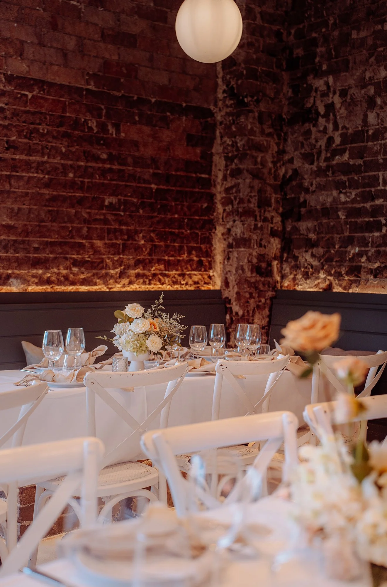 Elegant restaurant interior with a round table set with wine glasses, white napkins, and a floral centerpiece, against a backdrop of exposed brick walls and a ceiling-mounted spherical light.