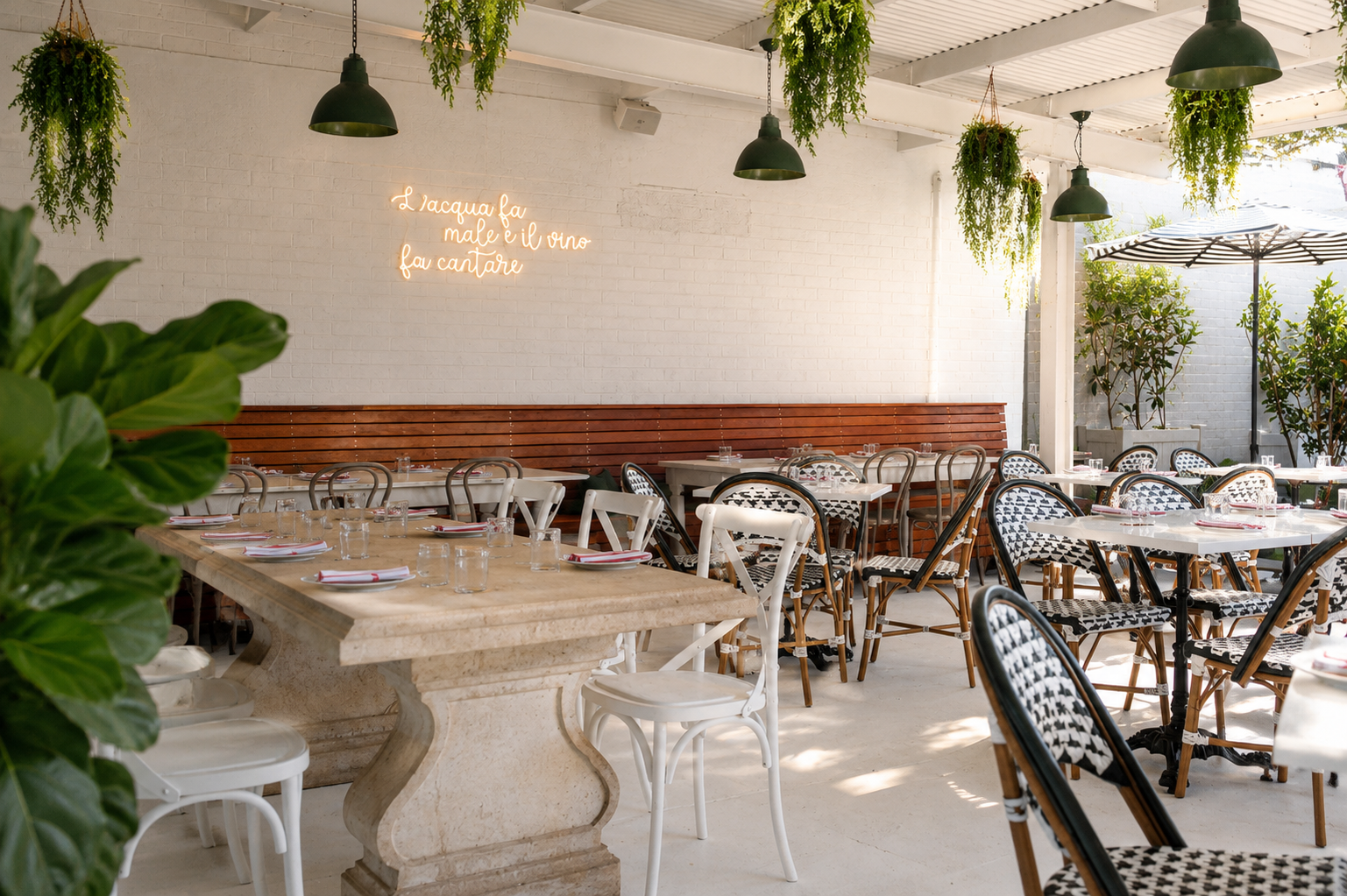 Outdoor restaurant patio with white brick walls, green plants hanging from the ceiling, and tables set with glassware and utensils. A neon sign on the wall reads in Italian: "L'acqua fa male e il vino fa cantare."