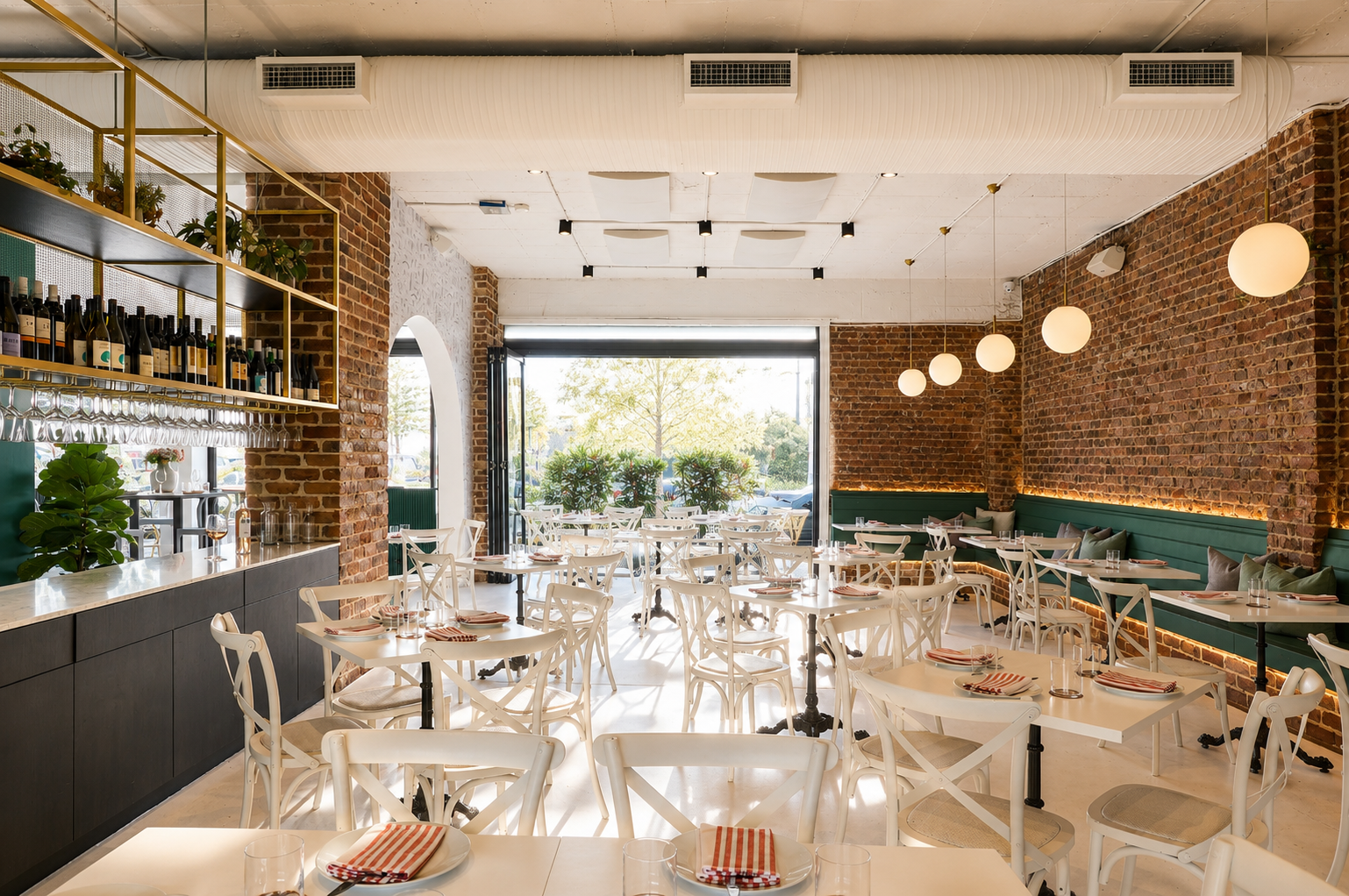 Bright restaurant interior with brick walls, white tables and chairs, green bench seating, hanging globe pendant lights, and large windows showing outdoor greenery.