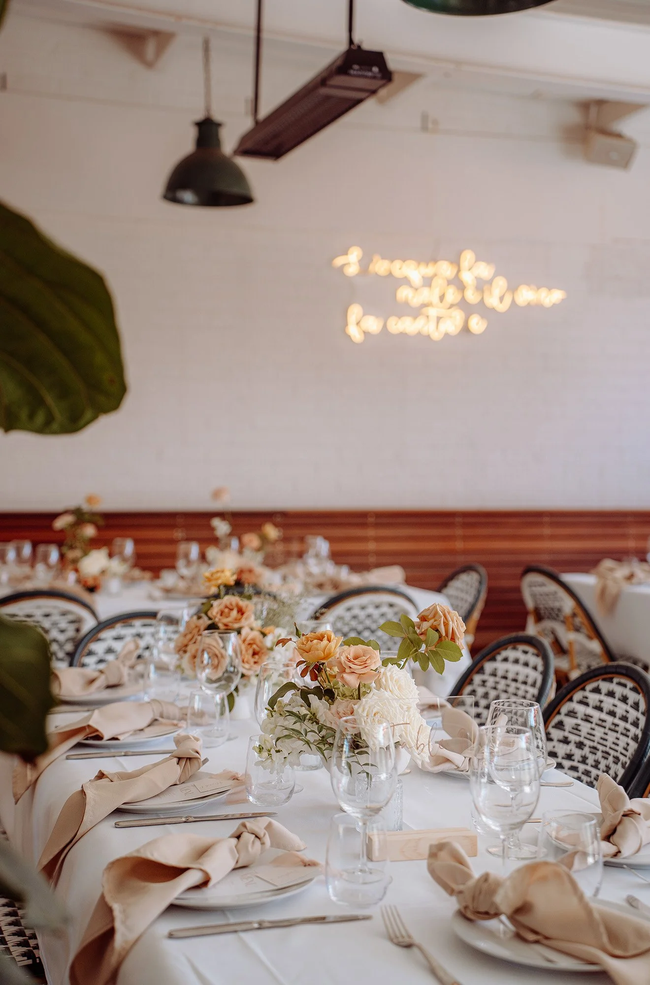 Elegant banquet table decorated with peach roses in clear glass vases, beige napkins tied with ribbons, and wine glasses, set in a decorated venue with flower arrangements.