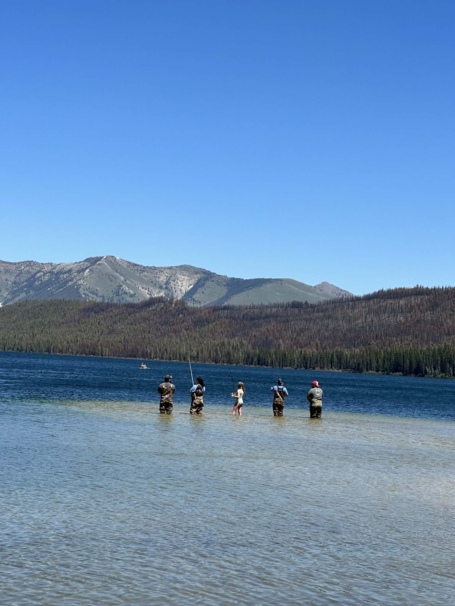 Anglers fly fishing in shallow water beside a scenic mountain lake under a clear blue sky.