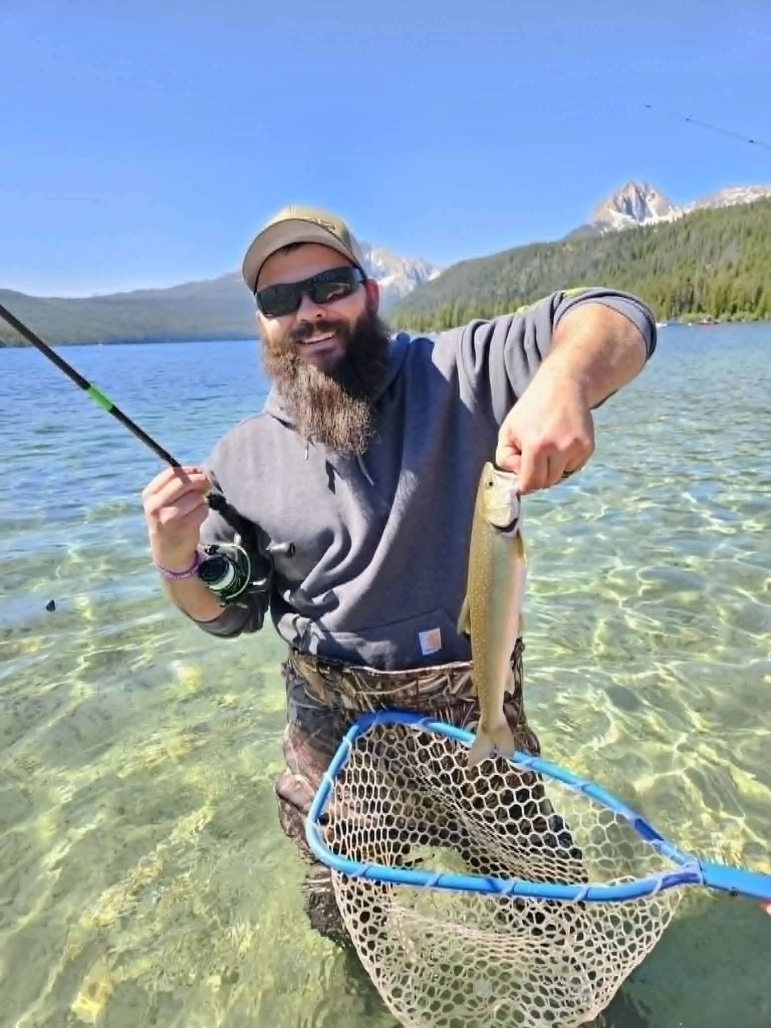 Man holding a fish with a fishing net in clear water, mountains in the background.