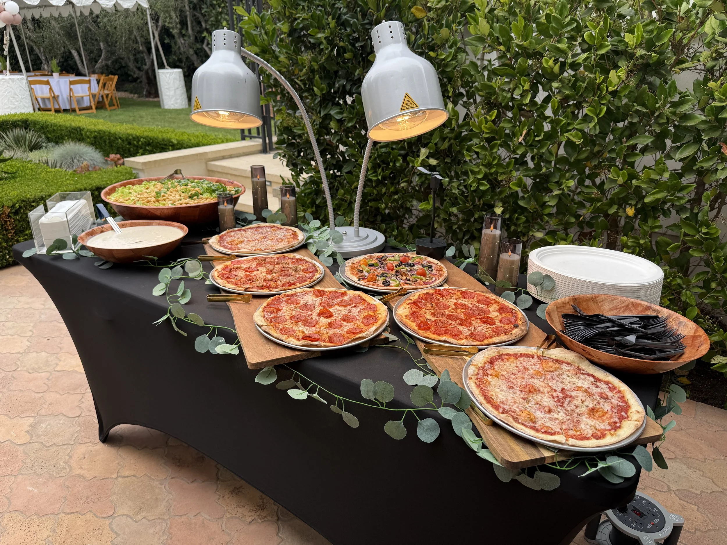 A buffet table with a black tablecloth displaying various pizzas, salad, and dipping sauces under white heat lamps, set outdoors with green bushes and a dining area in the background.