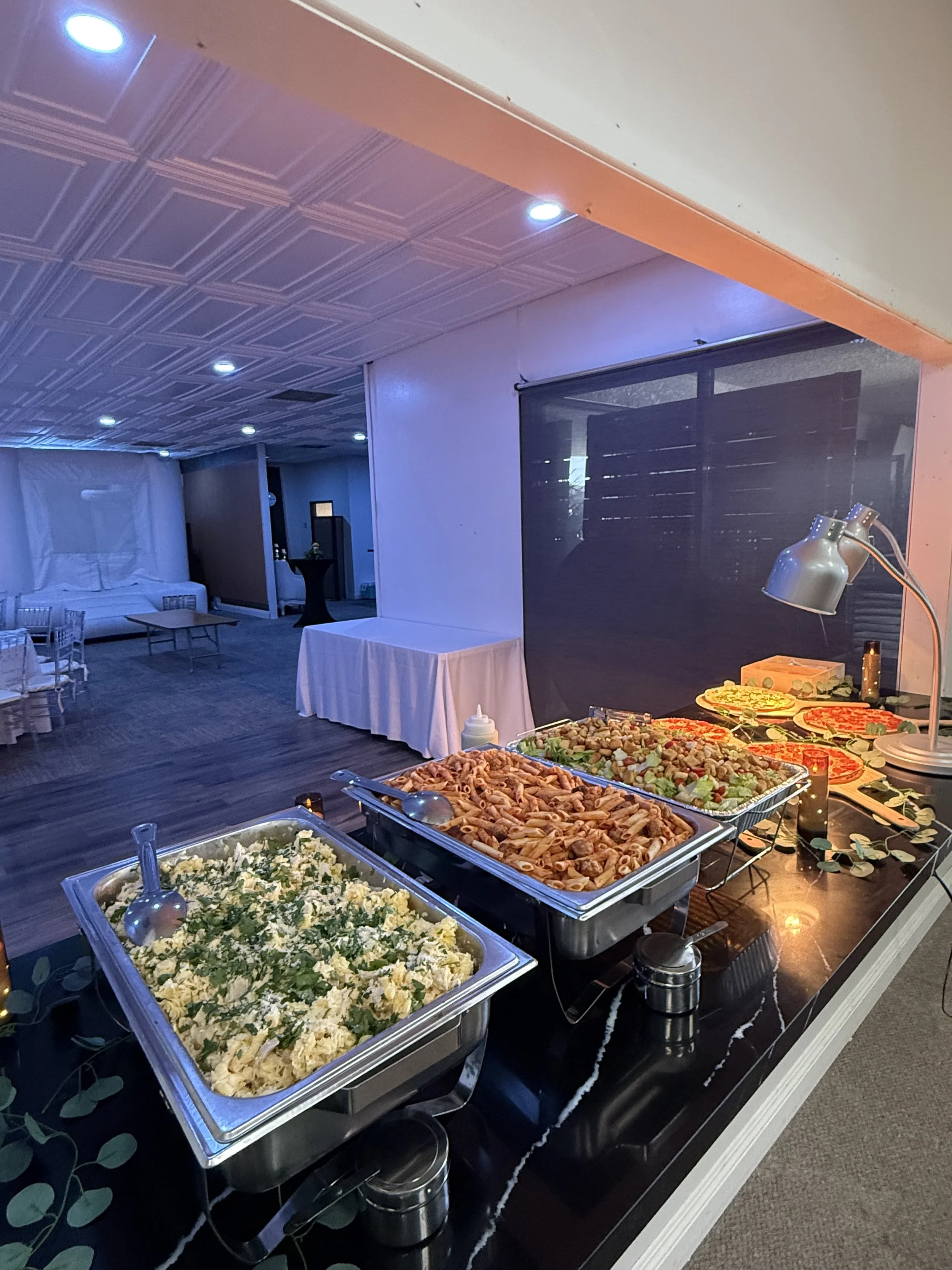 Buffet table with trays of pasta, salad, pizza, and other dishes set up in a decorated room with ceiling lights and seating area in the background.