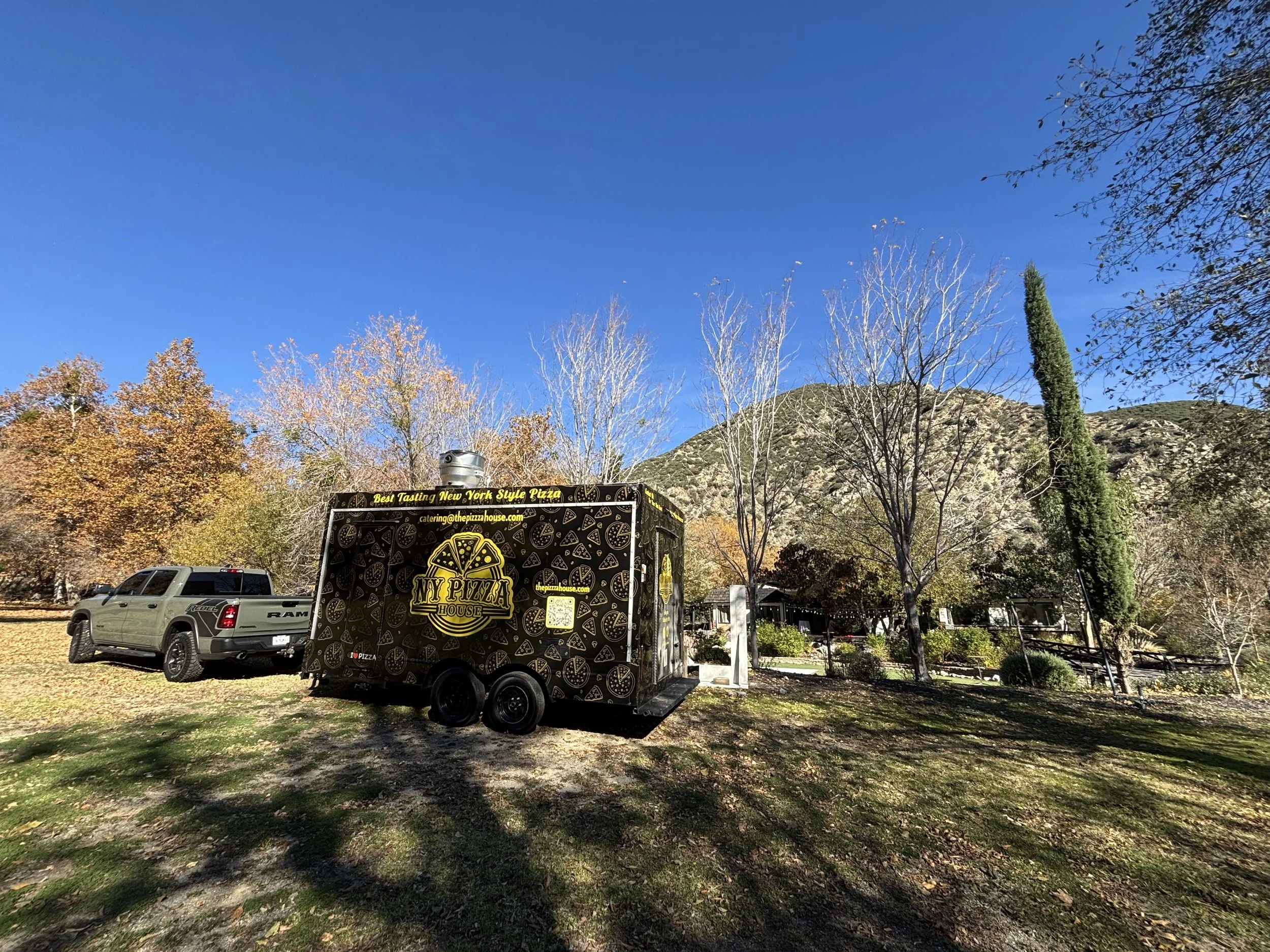 A black pizza delivery trailer with yellow lettering and pizza icons parked on a grassy area, with a white truck nearby, between trees with autumn foliage and a mountain in the background under a clear blue sky.