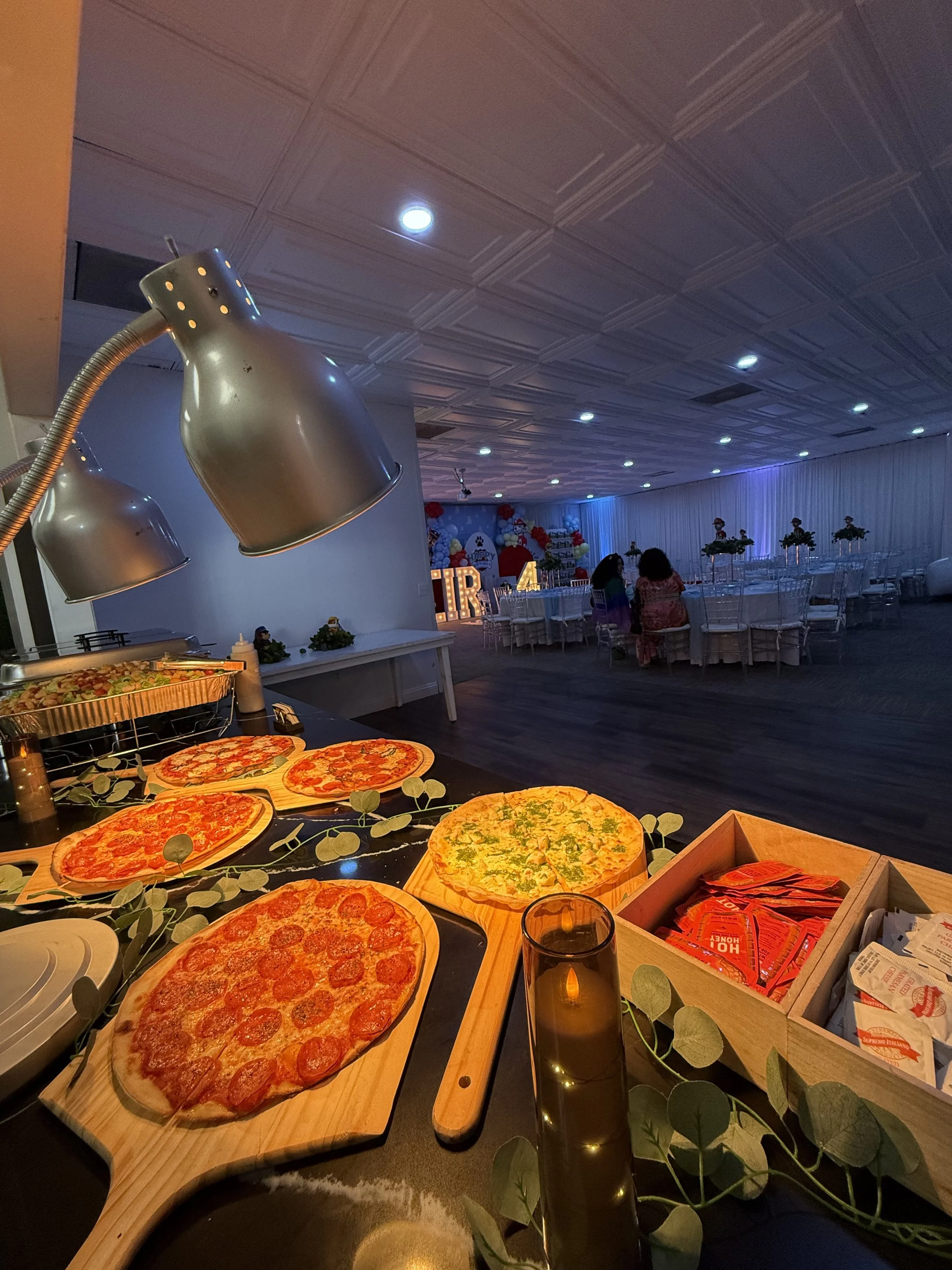 A buffet table with several pizza pizzas in front of a decorated party room with tables and chairs, balloons, and lights for a celebration.