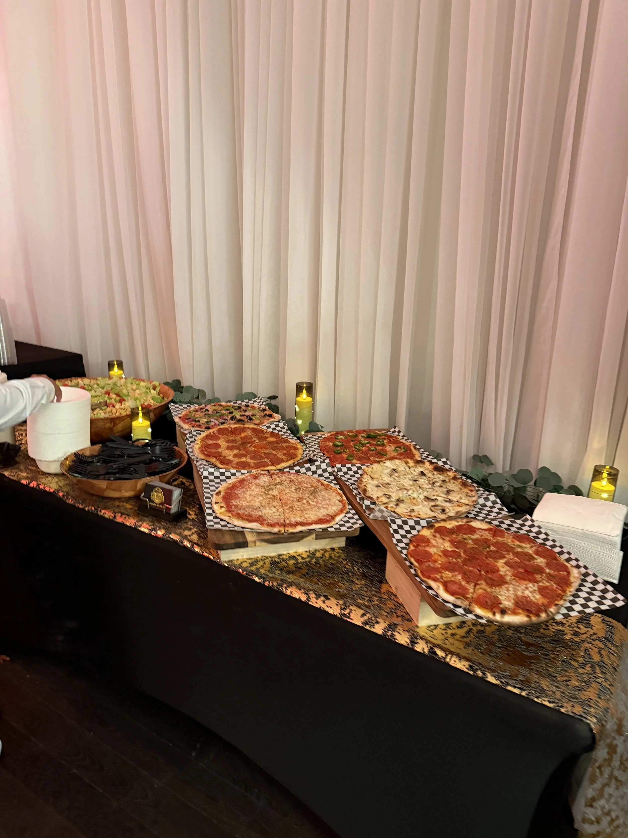 A table with various pizzas, salads, napkins, and candles on a checkered tablecloth, against a backdrop of white curtains.