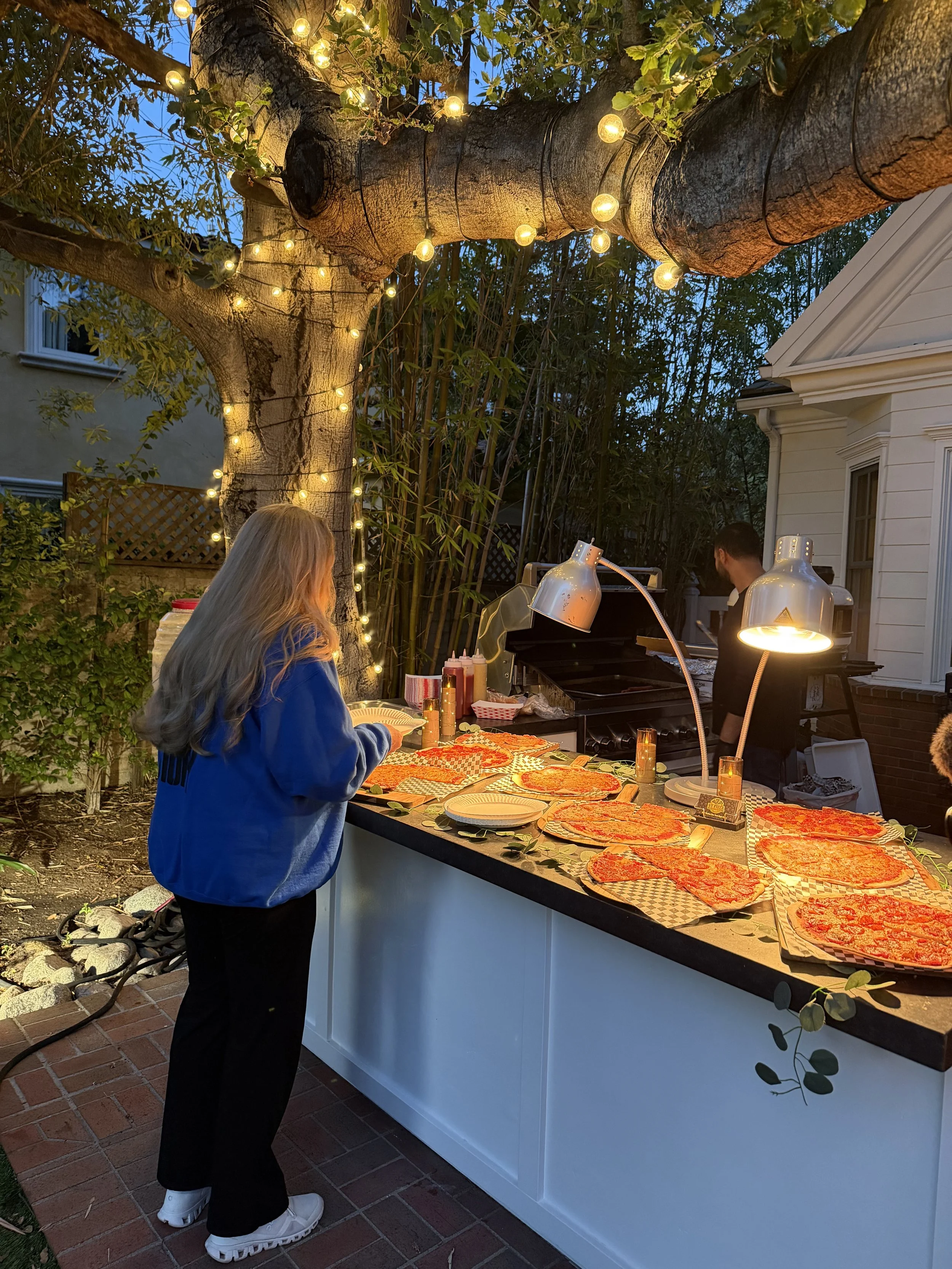A person with long blonde hair wearing a blue jacket and black pants stands at a pizza buffet table outdoors during evening. The table has several uncooked pizzas and condiments. There is a large tree decorated with string lights illuminated against 