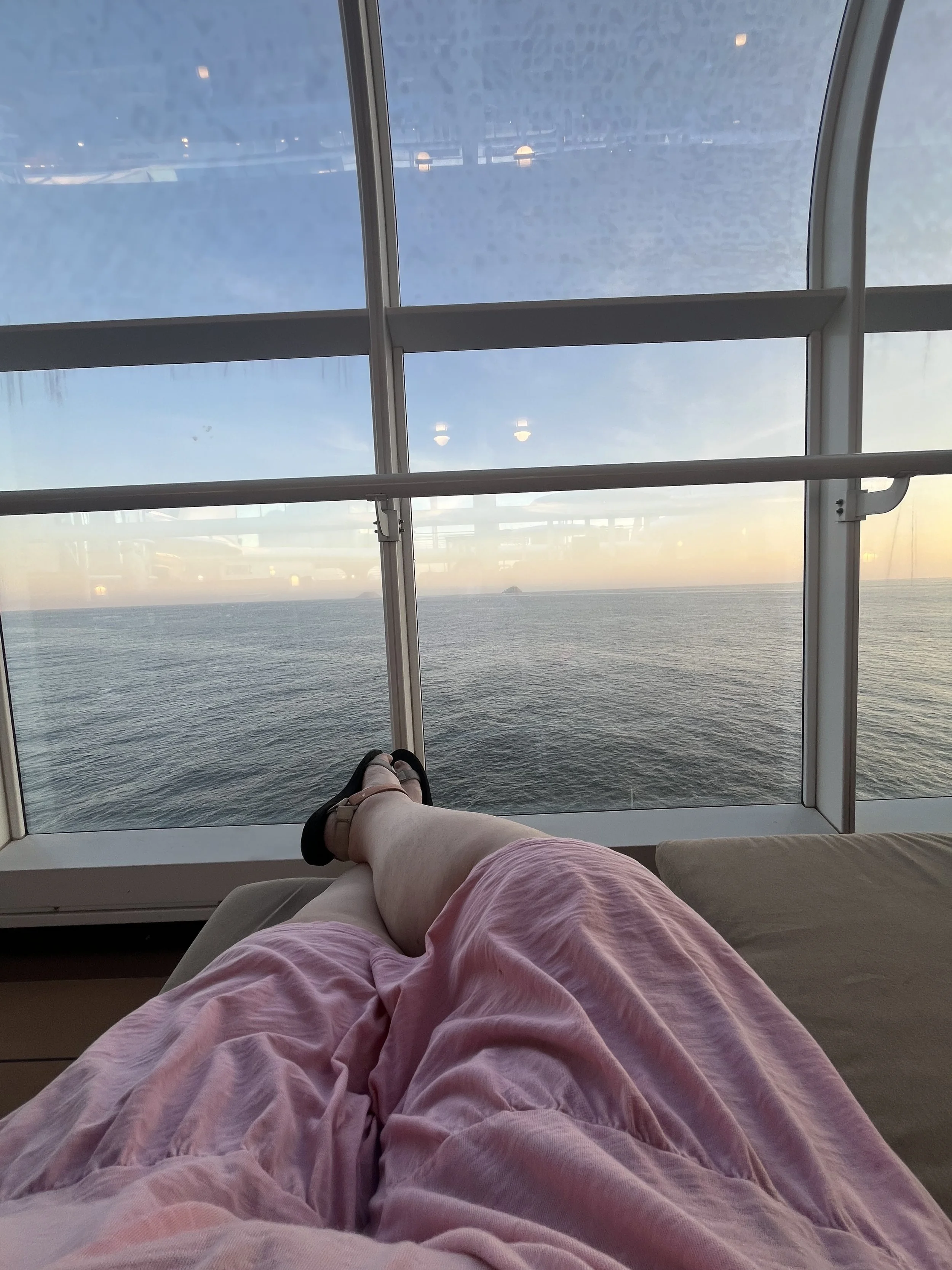 Woman in pink dress sitting a on sun lounger looking out over ocean on a cruise ship. Photo shows her legs on the lounger.