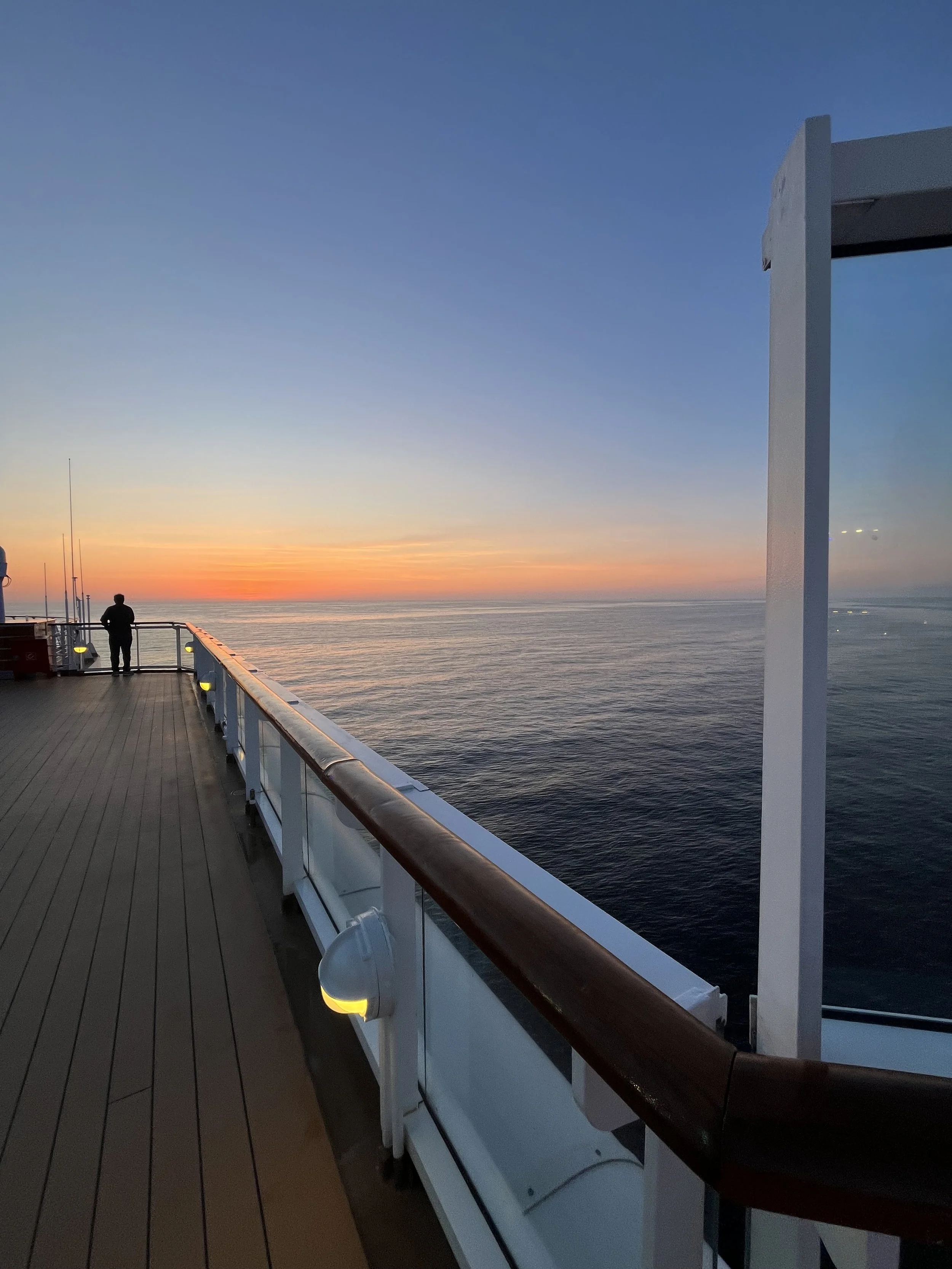 A person standing on the deck of a ship, looking at the sunset over the ocean, with clear skies and calm waters.