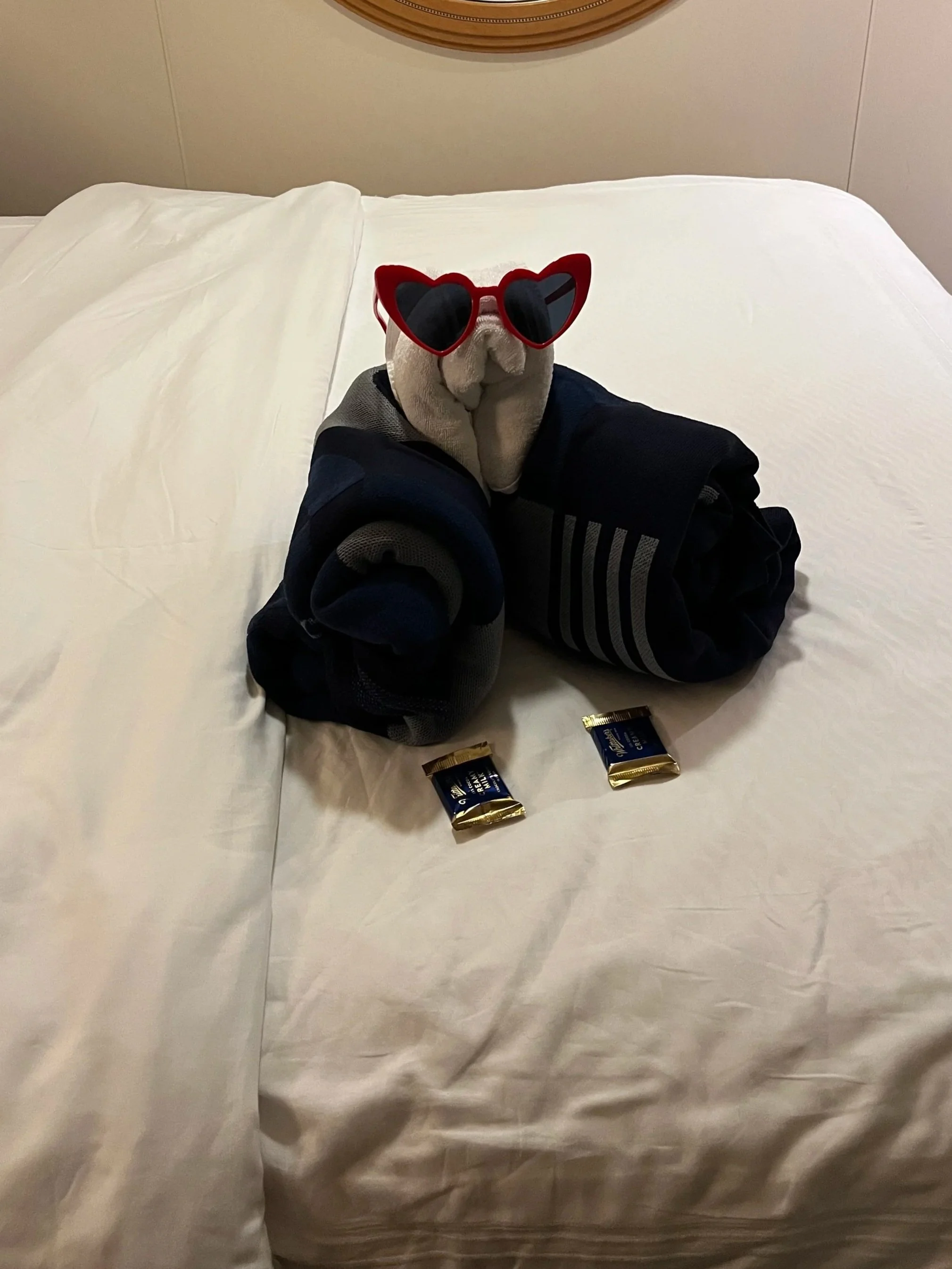Towel animal bear wearing heart shaped sunglasses on a white comforter with two chocolates sitting in front of it.