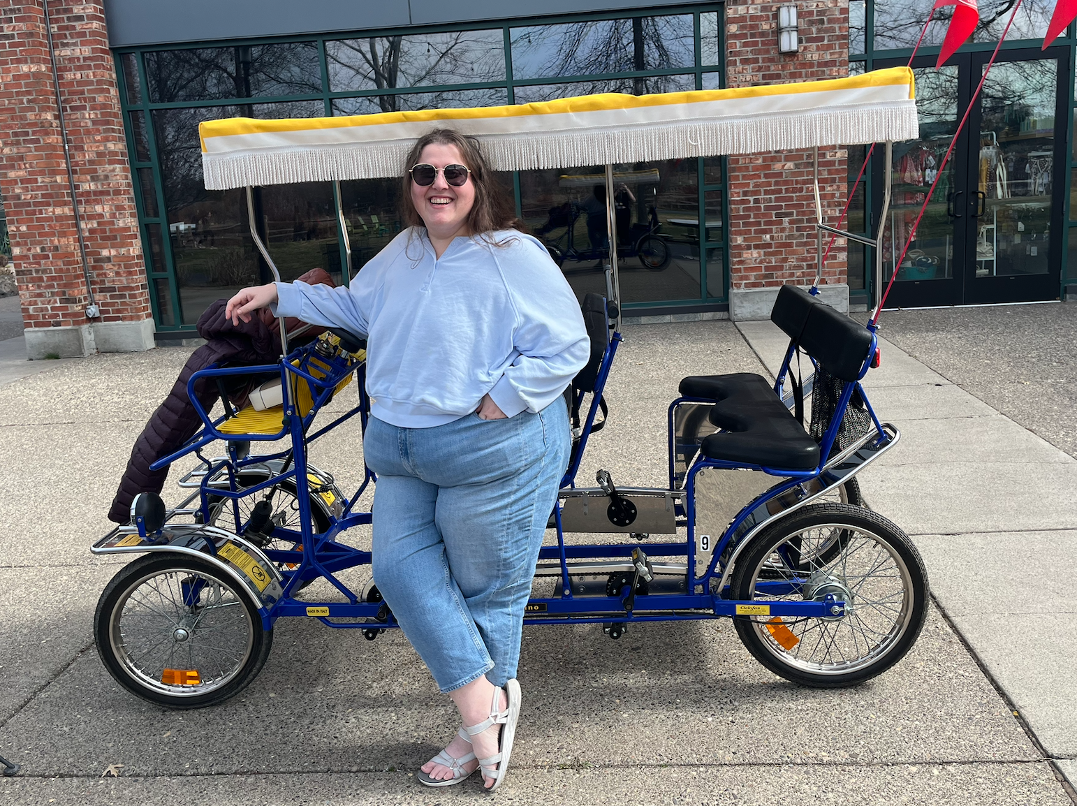 A woman in sunglasses smiling while standing next to a blue pedal-powered quadricycle with a canopy outside a building with brick and glass windows.