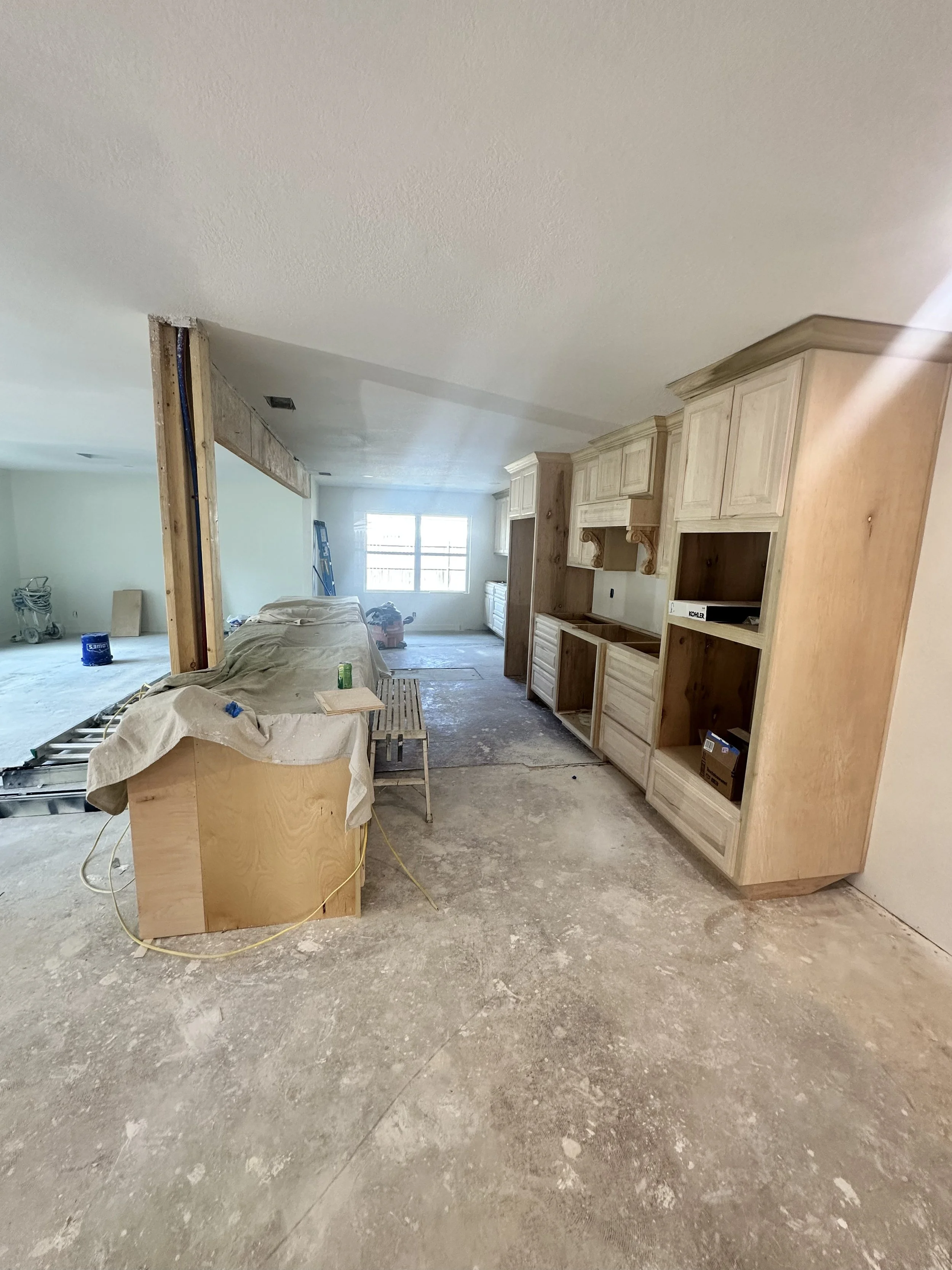 Interior of a house under renovation, with unfinished flooring, wooden cabinetry, construction tools, and materials scattered around.