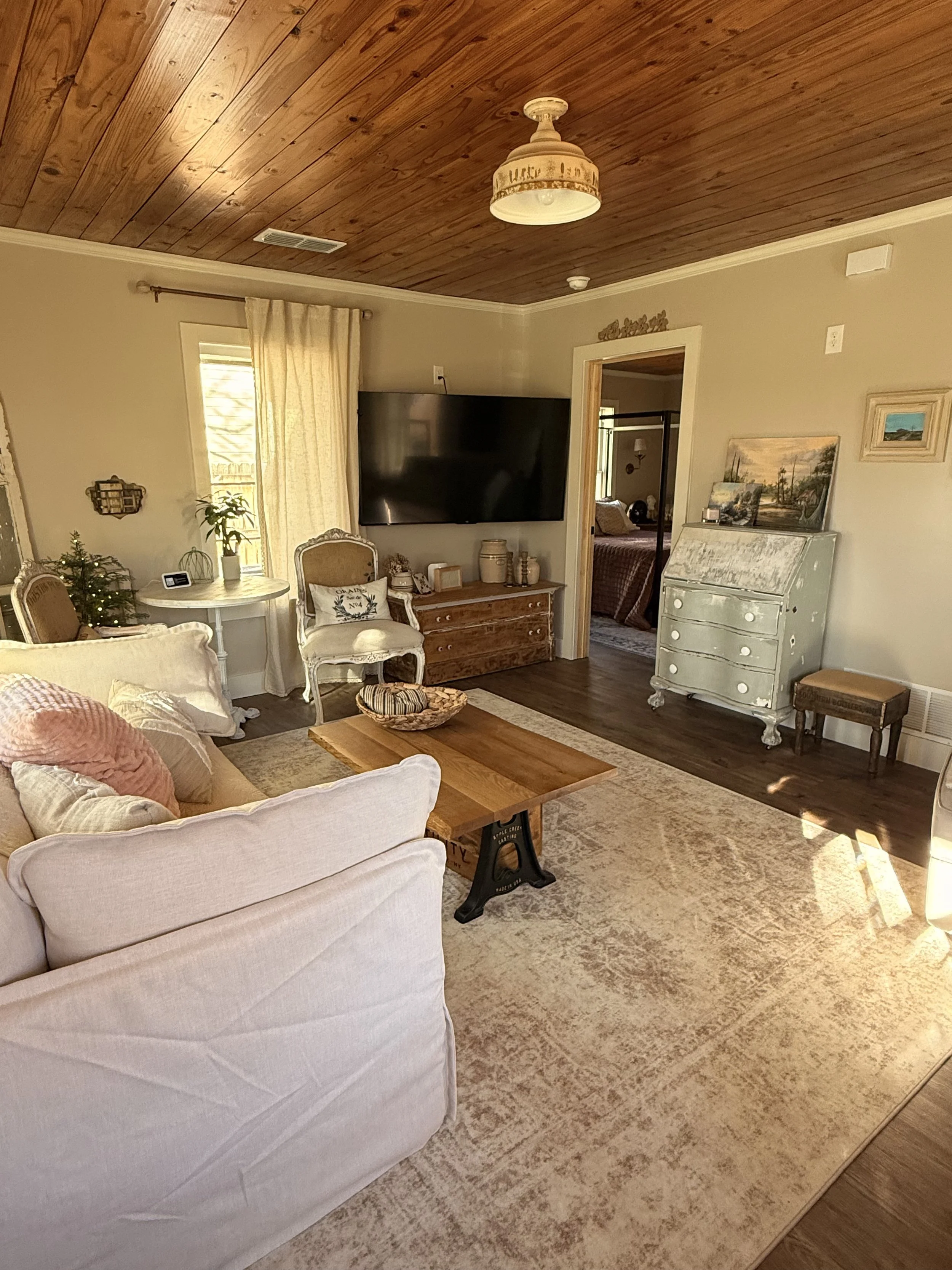 A living room with a beige sofa, a round white side table with a small Christmas tree, armchairs, a wooden coffee table, a flat-screen TV on the wall, various decorative items, and a doorway leading to a bedroom with a bed and window. The room has a wooden ceiling and a beige rug on hardwood floors.