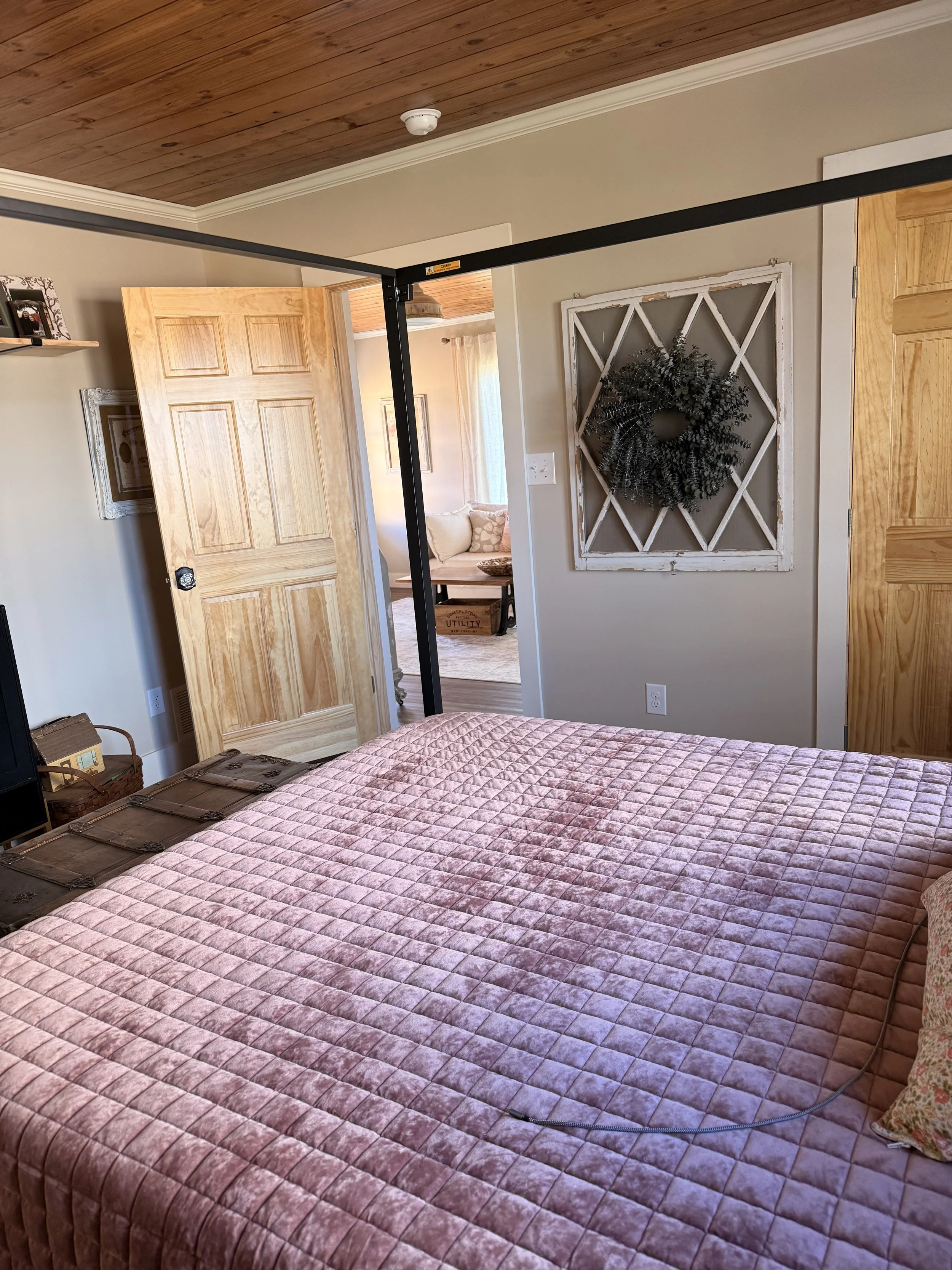 A bedroom with a pink quilted bedspread, wooden ceiling, open wooden door, wall decor featuring a circular wreath, and a glimpse of a sitting area with a beige couch in the background.