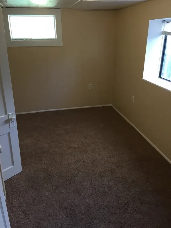 Empty beige-colored room with brown carpet flooring, two small windows with white frames, and a white door partially open.