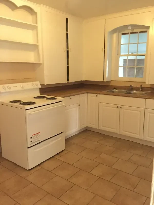Empty kitchen with white cabinets, a stove, a dark countertop, a double sink, and a window.