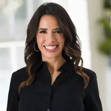 A smiling woman with dark brown hair styled in loose waves, wearing a black button-up shirt, hoop earrings, and in a bright, modern interior setting.