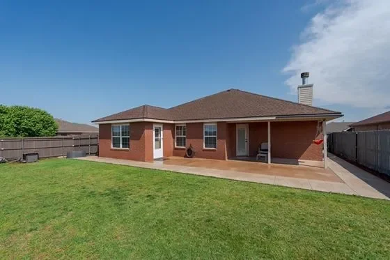 Backyard of a brick house with a green lawn, patio, and wooden fence under a blue sky.