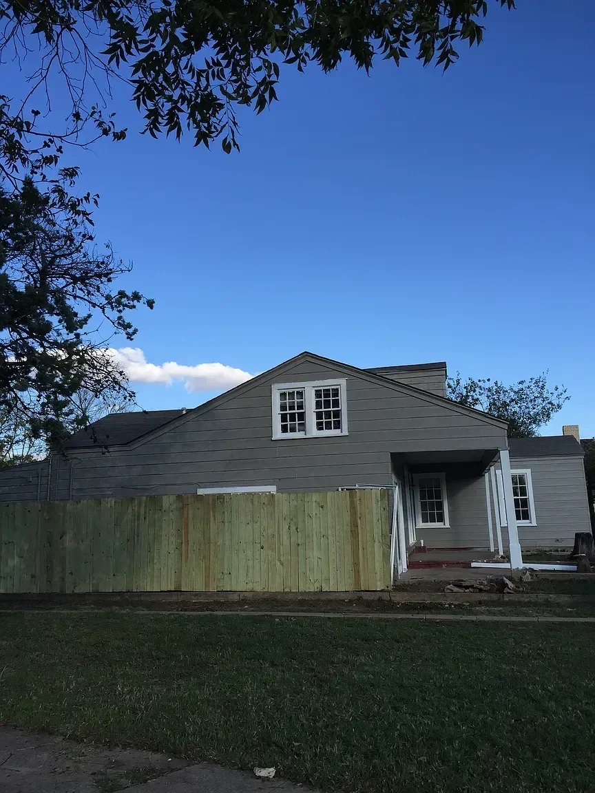 A house under construction with a wooden privacy fence in front, gray siding, white windows, and a small porch, set against a blue sky with a few clouds.