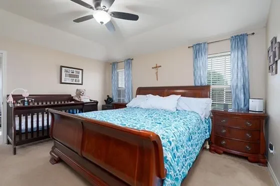 Bedroom with wooden bed, cross on wall, ceiling fan, blue curtains, window blinds, dresser, nightstand, and crib.