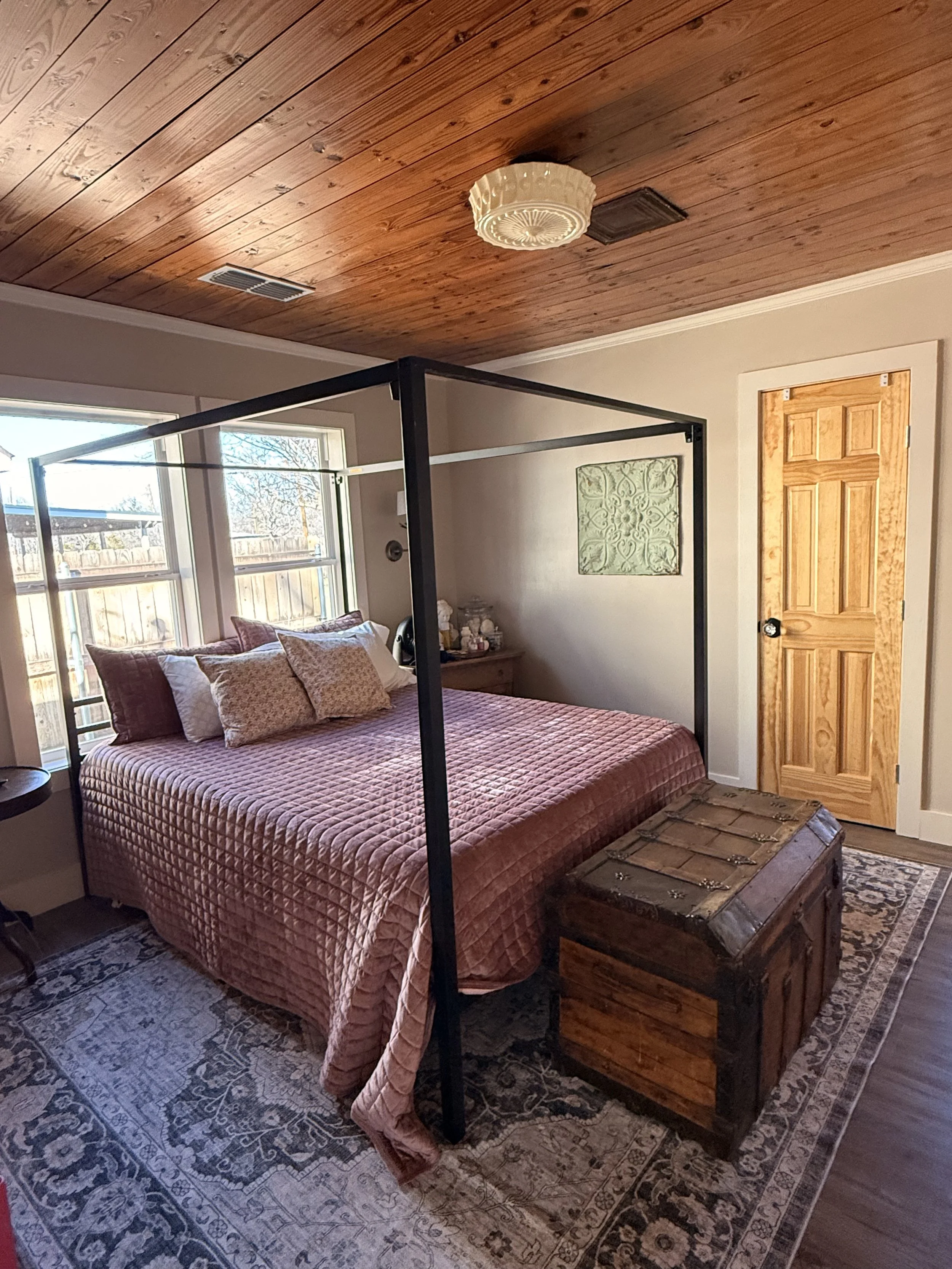 A bedroom with a four-poster bed, pink quilt, and patterned pillows, a wooden chest at the foot, large windows with views outside, and a wooden ceiling with a ceiling light and vents.