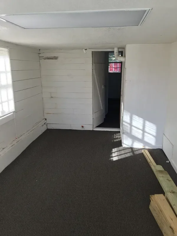 Empty room with white painted wooden walls, a dark carpeted floor, and a small door opening into a dark space. Sunlight casts shadows on the floor from a window outside the room. Construction wooden planks are on the right side of the room.