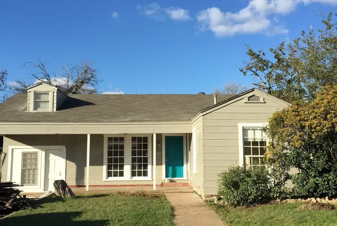 A house with gray siding, white trim, and a blue front door under a partly cloudy sky, with trees and shrubs in the yard.