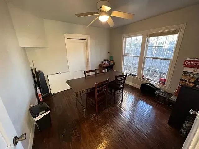 Dining room with wooden table and four chairs, ceiling fan with light, two large windows, and a door. Items include a black speaker, a small table with toys or games, and a trash bin.