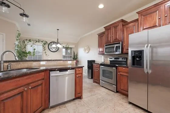 Modern kitchen with wooden cabinets, stainless steel appliances, granite countertops, tiled floor, and a view into the dining area with windows and a clock.