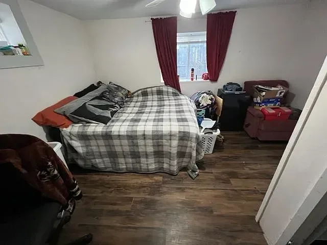 A bedroom with a bed covered in a plaid blanket, pillows, and various items on it, a window with red curtains, and a reddish sitting chair with boxes and clothes nearby.