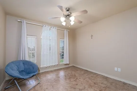 Empty room with beige walls, three windows with white curtains, a blue papasan chair, a ceiling fan with lights, and a small wall cross.