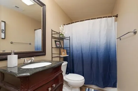 A bathroom with a dark granite countertop vanity, a mirror with a silver frame, a toilet, a blue and white ombre shower curtain, a metal shelving unit with decorative items, and a towel bar on the wall.