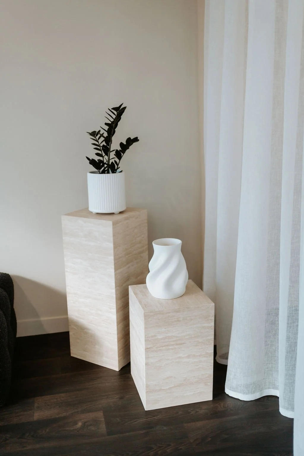 A black plant with long, wide leaves in a white textured pot on a beige wooden pedestal, and a white abstract ceramic vase on a smaller beige wooden pedestal, with a background of a beige wall and sheer white curtains.