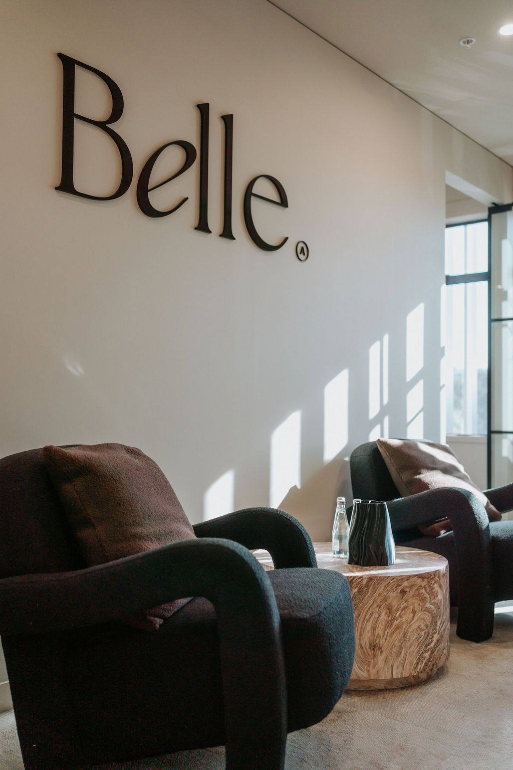 Modern lobby with two black armchairs, a round wooden coffee table, and sunlight casting shadows on a white wall with the word "Belle" mounted on it.