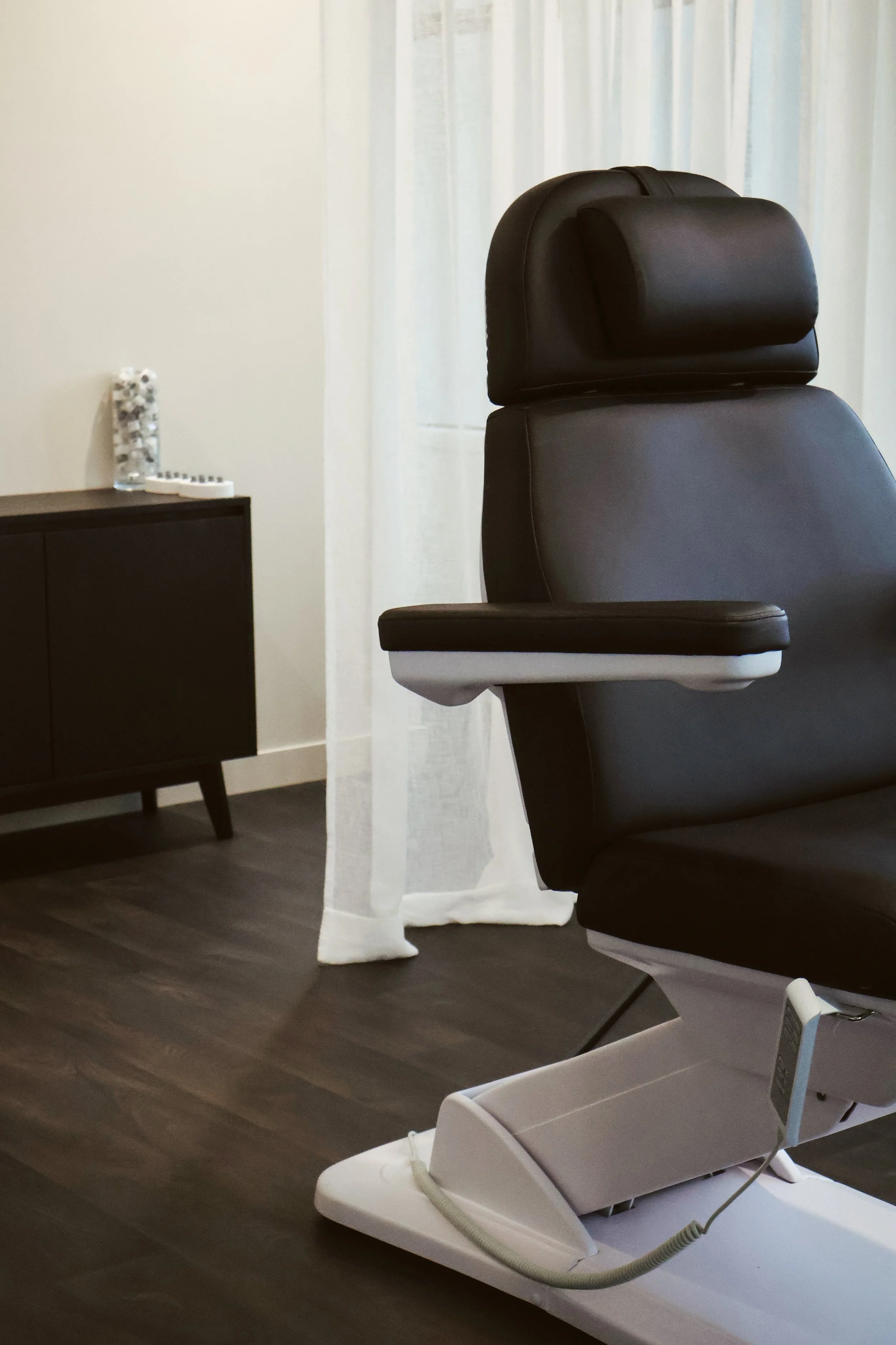A black massage chair in a room with white curtains, a dark wooden floor, a black side table with a jar of stones and a white extension cord, and a white recliner with a cable attached.