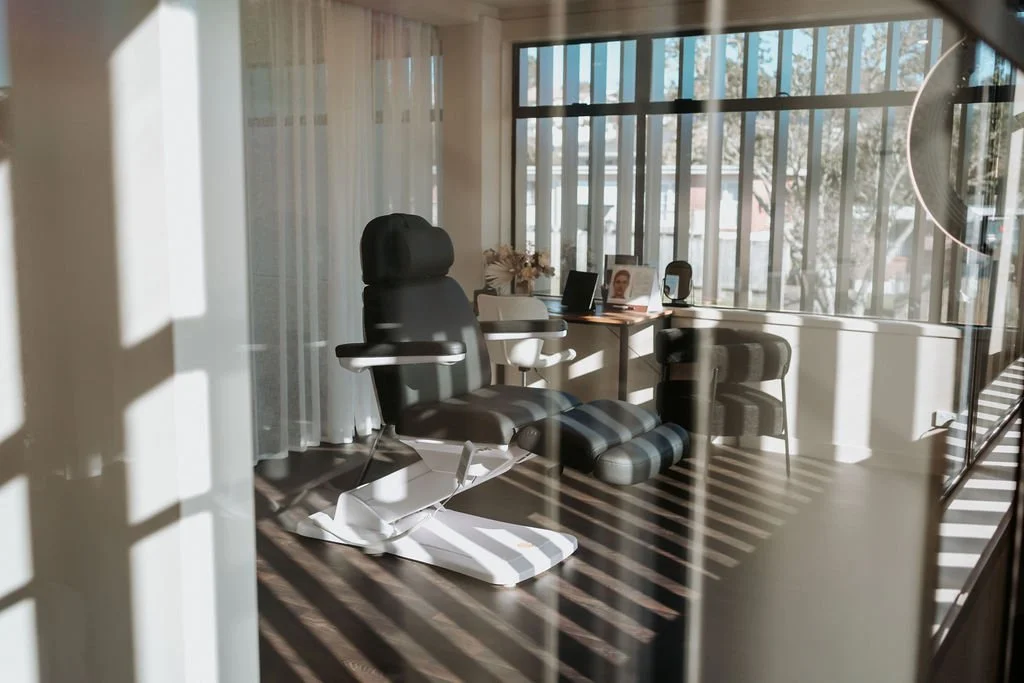 Medical examination room with an adjustable chair, a desk with photo frames, a phone, and flowers, illuminated by sunlight through large windows with blinds.