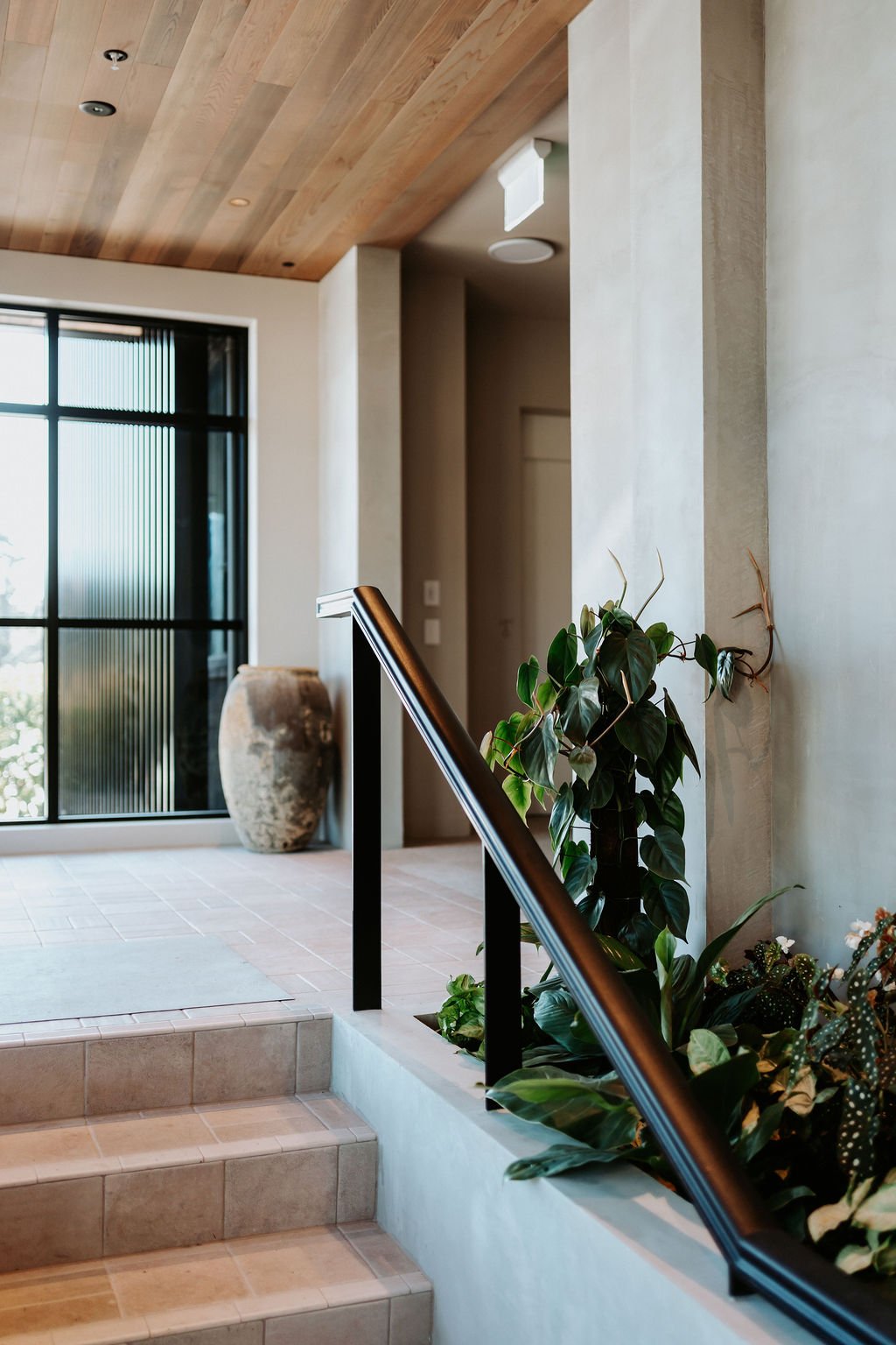 Interior of a modern home with tiled stairs, a potted plant, large window with vertical blinds, and a wooden ceiling.