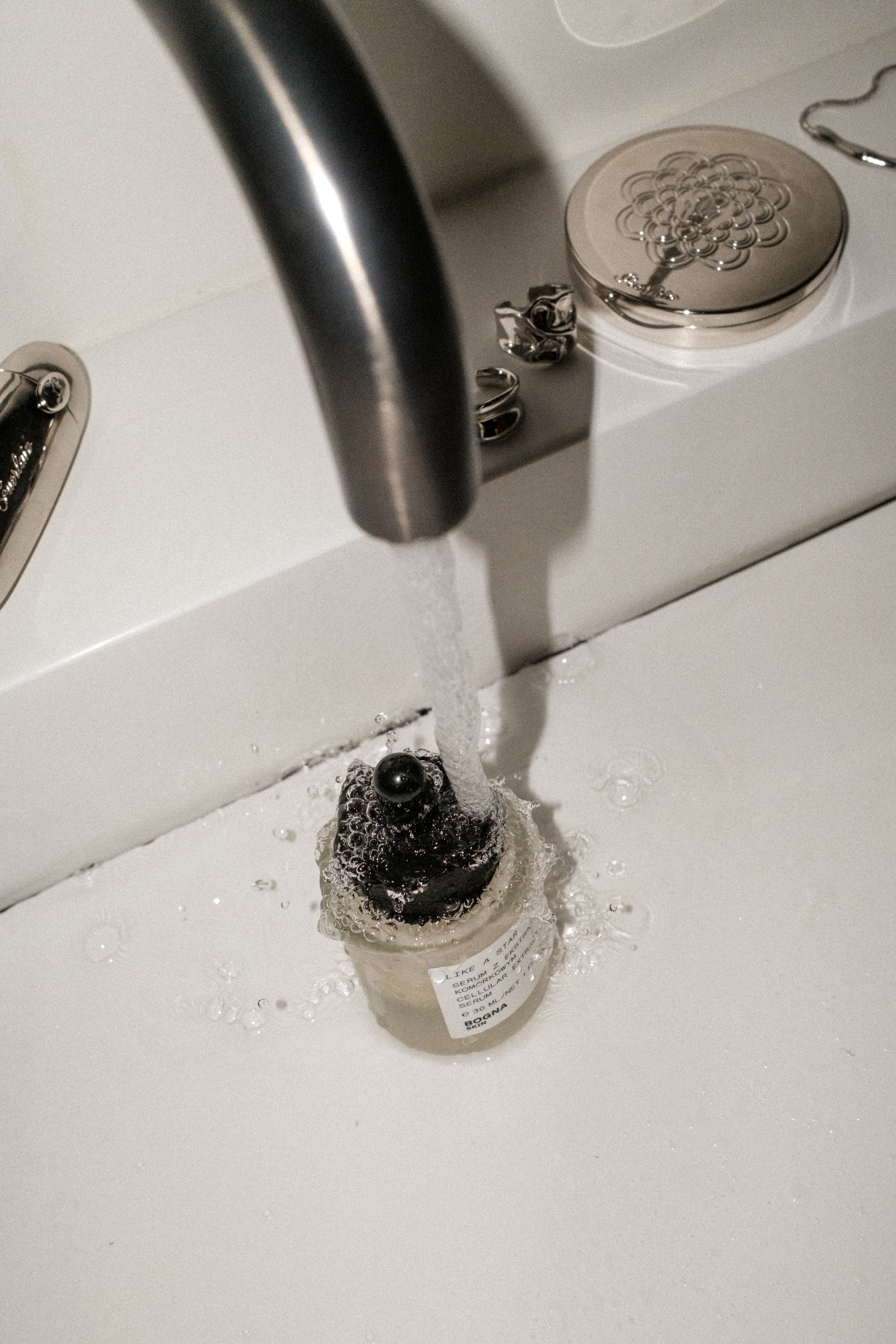 A small bottle with a dark liquid inside is placed in a white sink, with water running from the faucet onto the bottle. The bottle has a label with some text, and a small black ball is placed on top of the bottle. Silver jewelry is visible near the faucet.