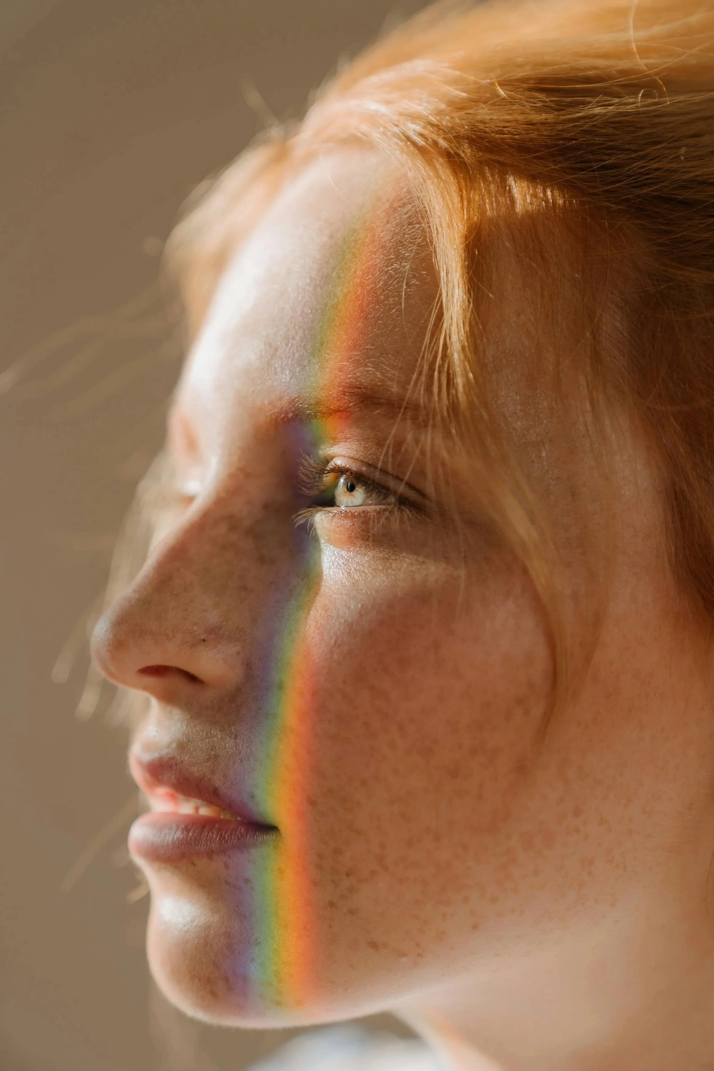 Close-up of a person's side face with light brown hair and freckles, featuring a rainbow reflection across their face from the left side to the right cheek.