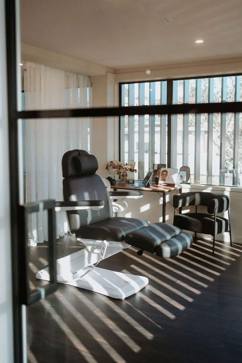 An office space seen through a glass door, featuring a black ergonomic chair, a small black and gray upholstered armchair, a wooden desk with various objects including a laptop and a framed photo, and a large window with vertical blinds and sheer curtains, casting striped shadows on the dark wooden floor.