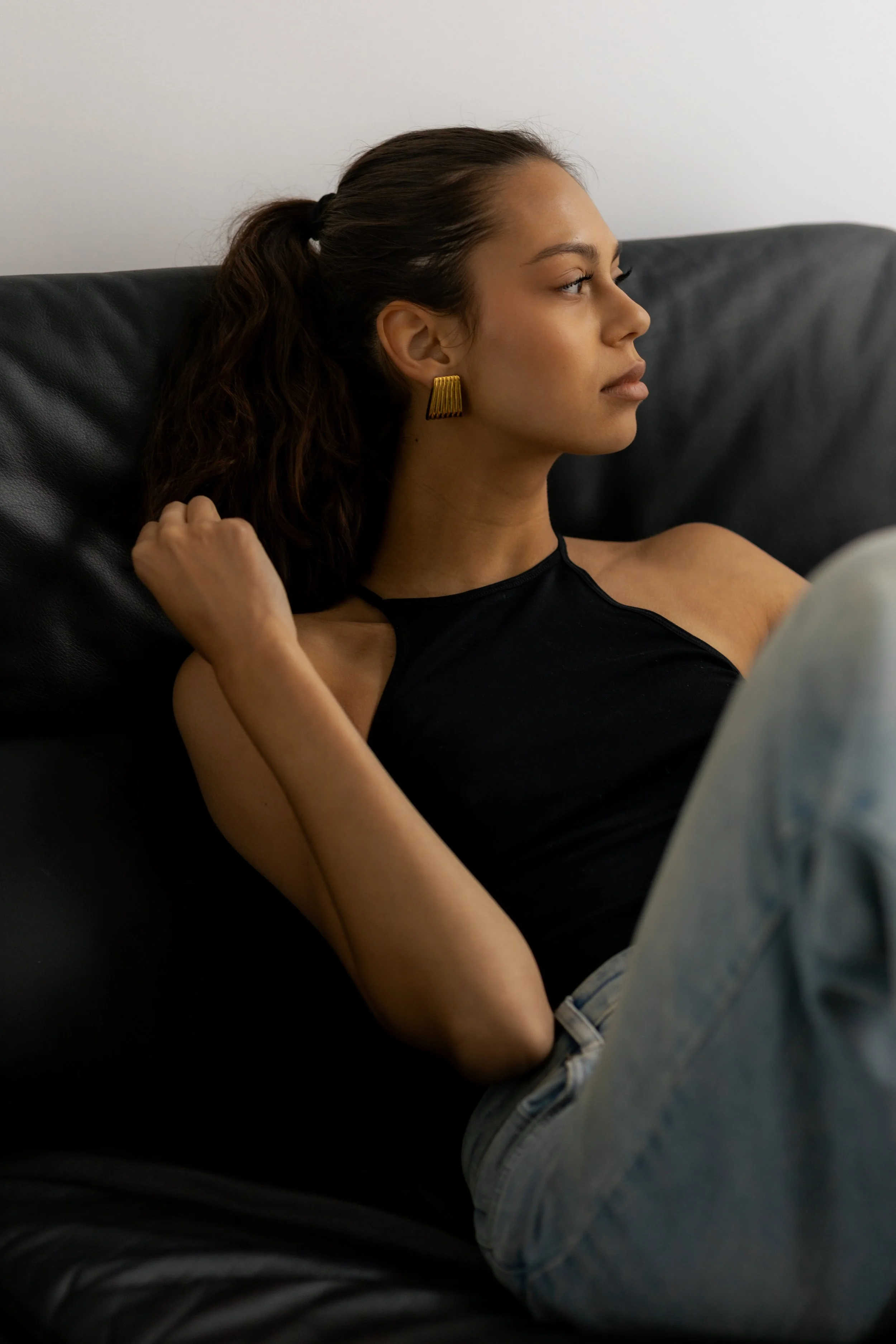 Side view of a young woman with dark hair pulled back in a ponytail, wearing gold earrings and a black tank top, sitting on a black leather sofa against a plain light-colored wall, looking to the right.