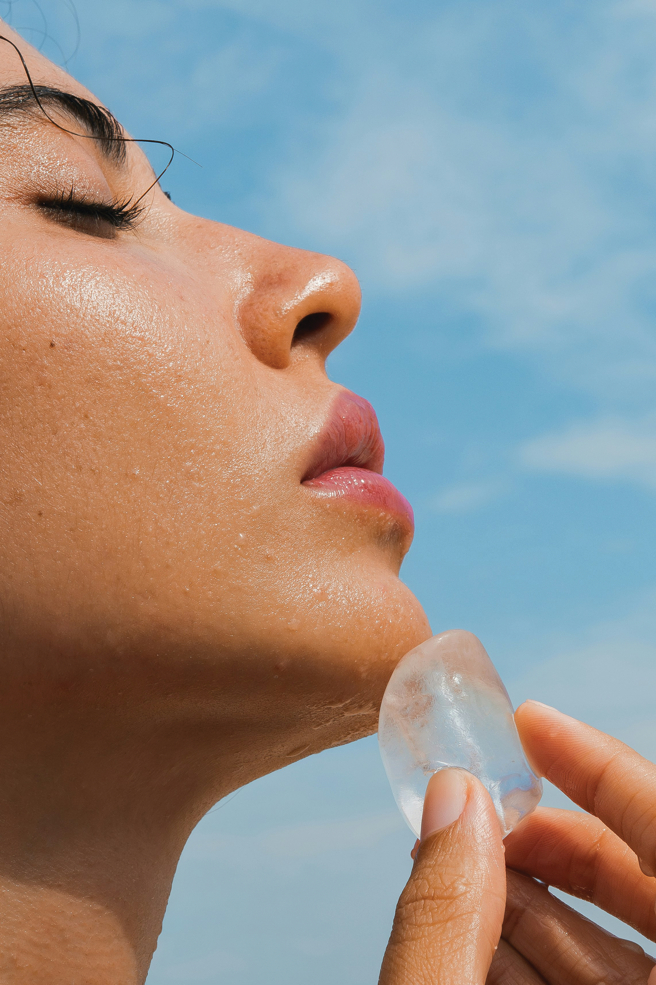 Close-up of a person's face with wet skin holding an ice cube near the chin, against a blue sky background.