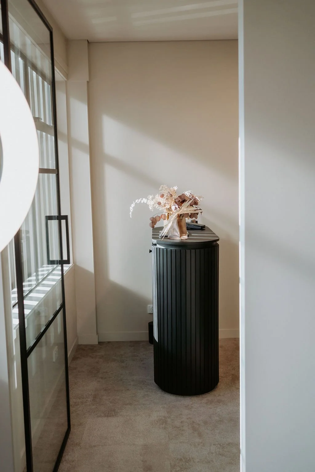 A small corner in a room with beige walls, a black vertical cabinet, and a dried flower arrangement on top, near a window with black framing.